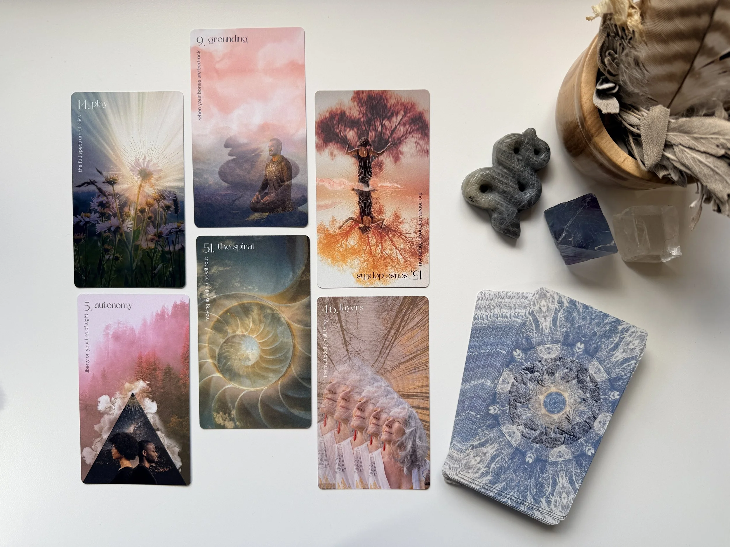 A spread of tarot or oracle cards with various artistic designs laid out on a white surface. Some cards depict natural landscapes, abstract patterns, and symbolic imagery. Nearby is a small stone sculpture and a container holding dried leaves or feathers.