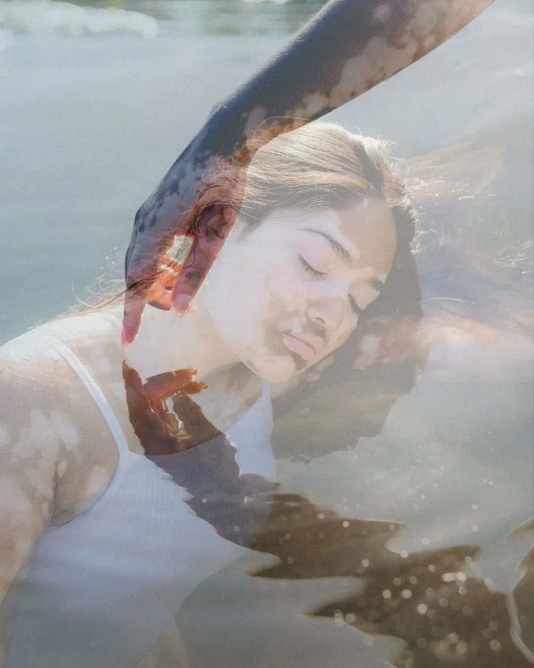 A woman with dark hair floating in water with her eyes closed, and a reflection of her face and arm in the water.