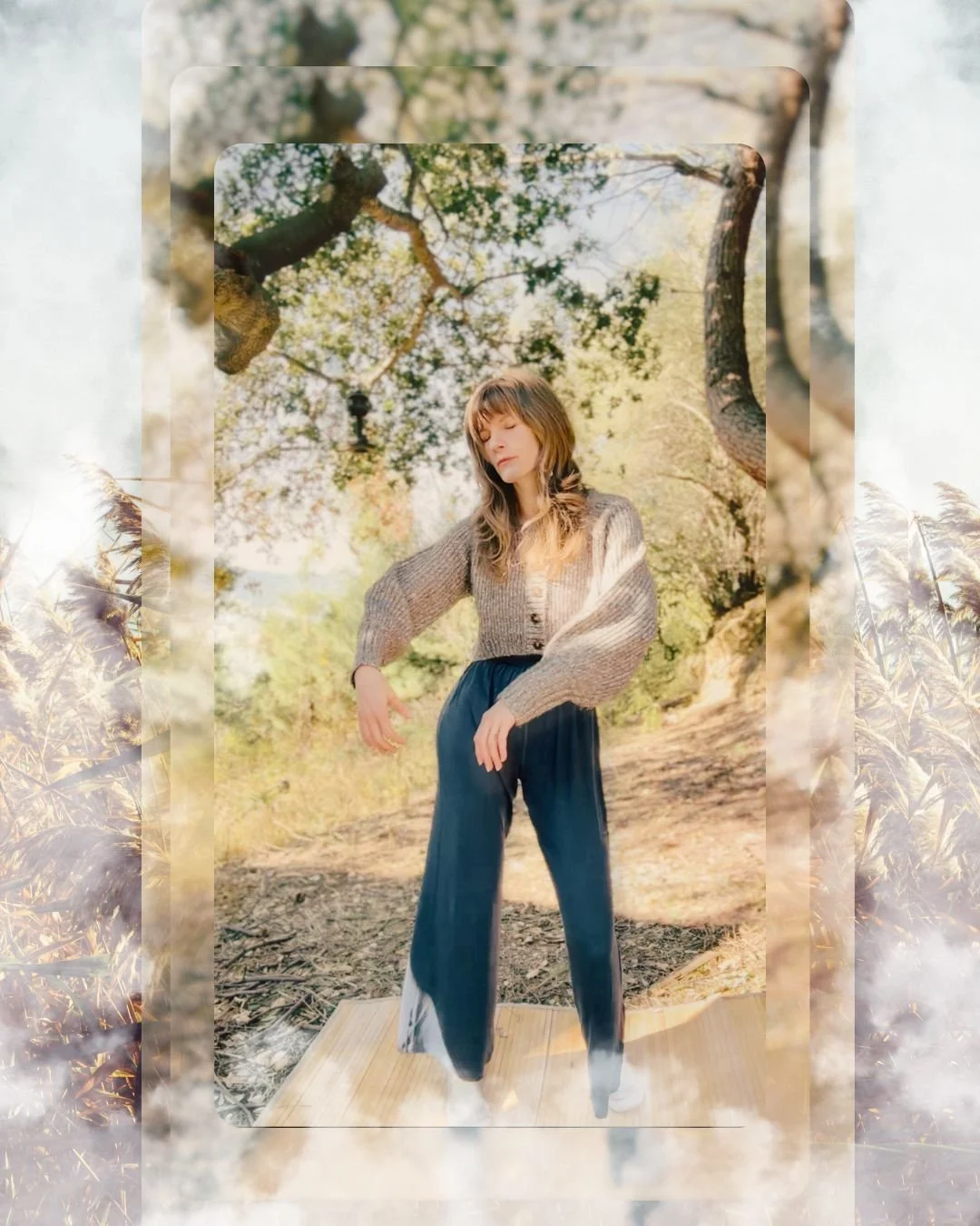 A woman with long hair and bangs, wearing a beige knitted cardigan and dark wide-leg pants, standing outdoors among trees with sunlight filtering through the branches.