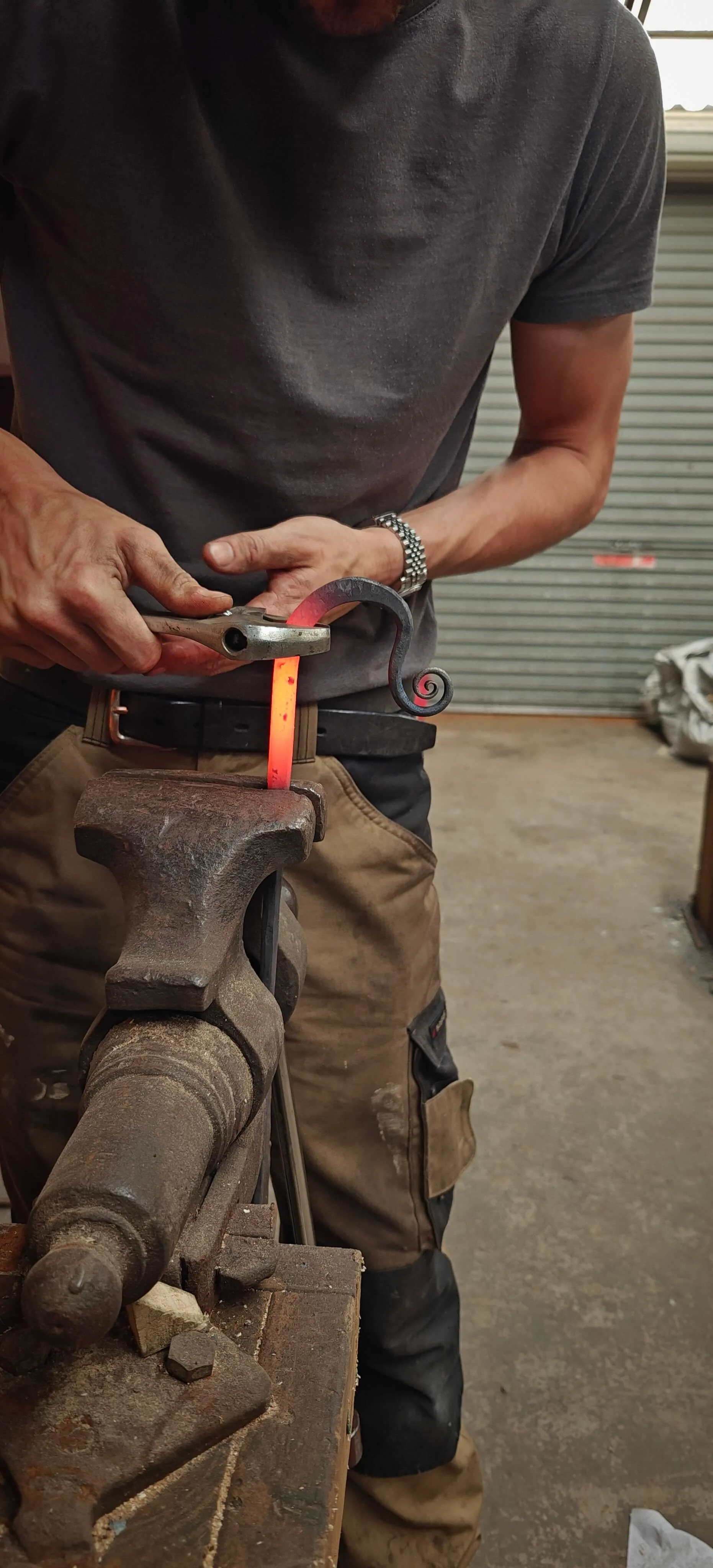 A blacksmith using his tools in Gabha na Lámh Fada (Long Arm Forge) to heat and shape a metal piece over an anvil in a workshop.