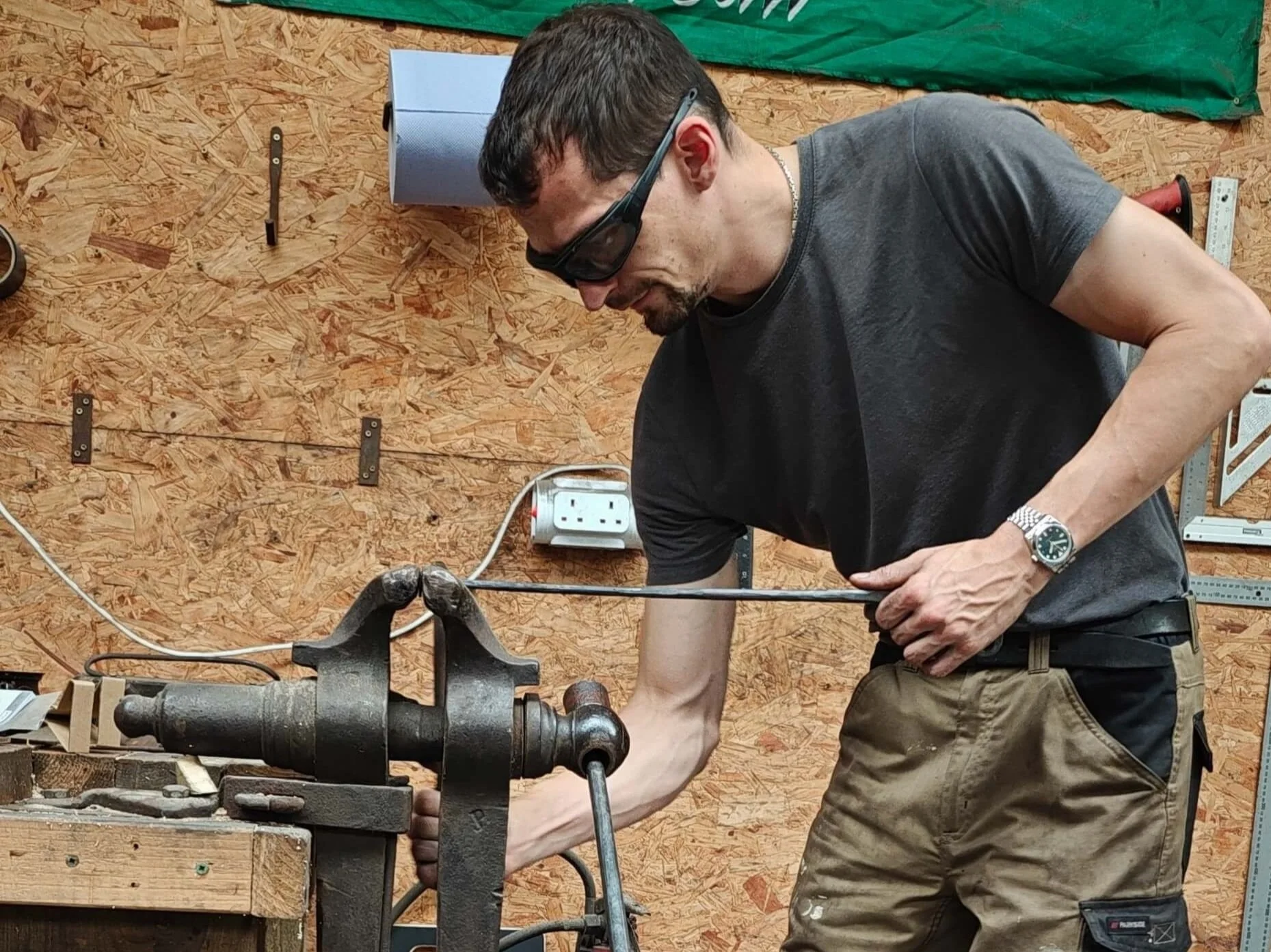 A blacksmith using a metal vice in Gabha na Lámh Fada (Long Arm Forge) while working on a piece of metalwork with various tools and wooden walls.