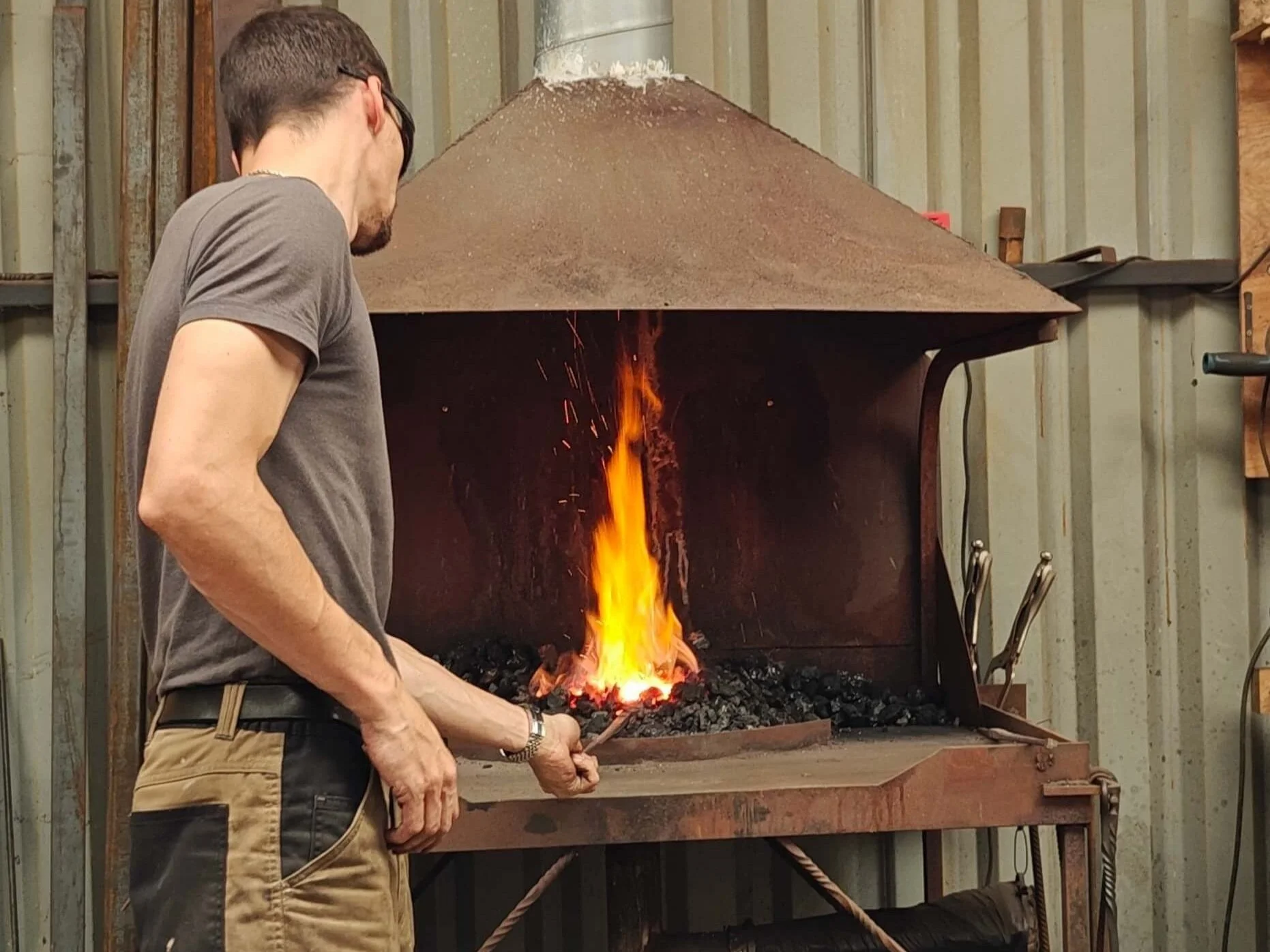 A blacksmith hand-forging metal in Gabha na Lámh Fada (Long Arm Forge), with a fire glowing inside a forge.