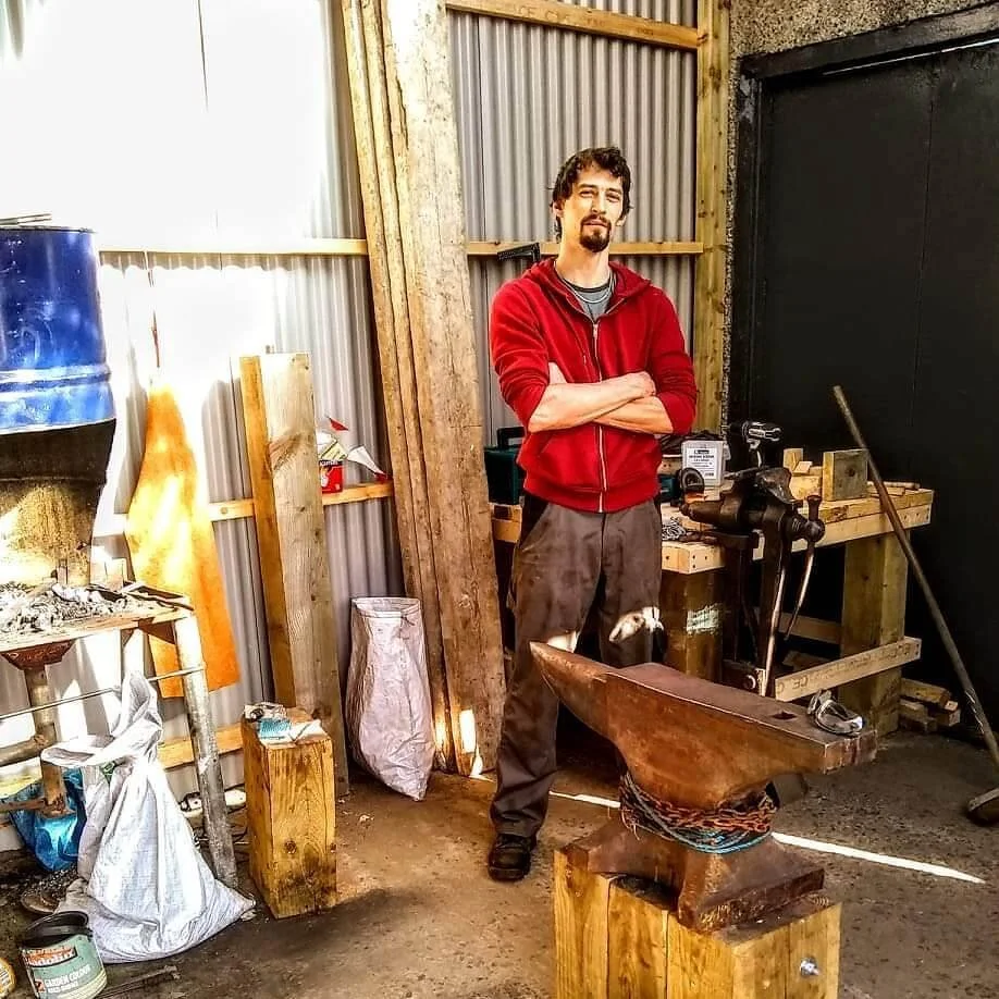 A blacksmith with dark hair and a beard stands with arms crossed in the workshop of Gabha na Lámh Fada (Long Arm Forge), wearing a red hoodie and brown work pants, with various metalwork tools, wooden planks, and an anvil in front of him.