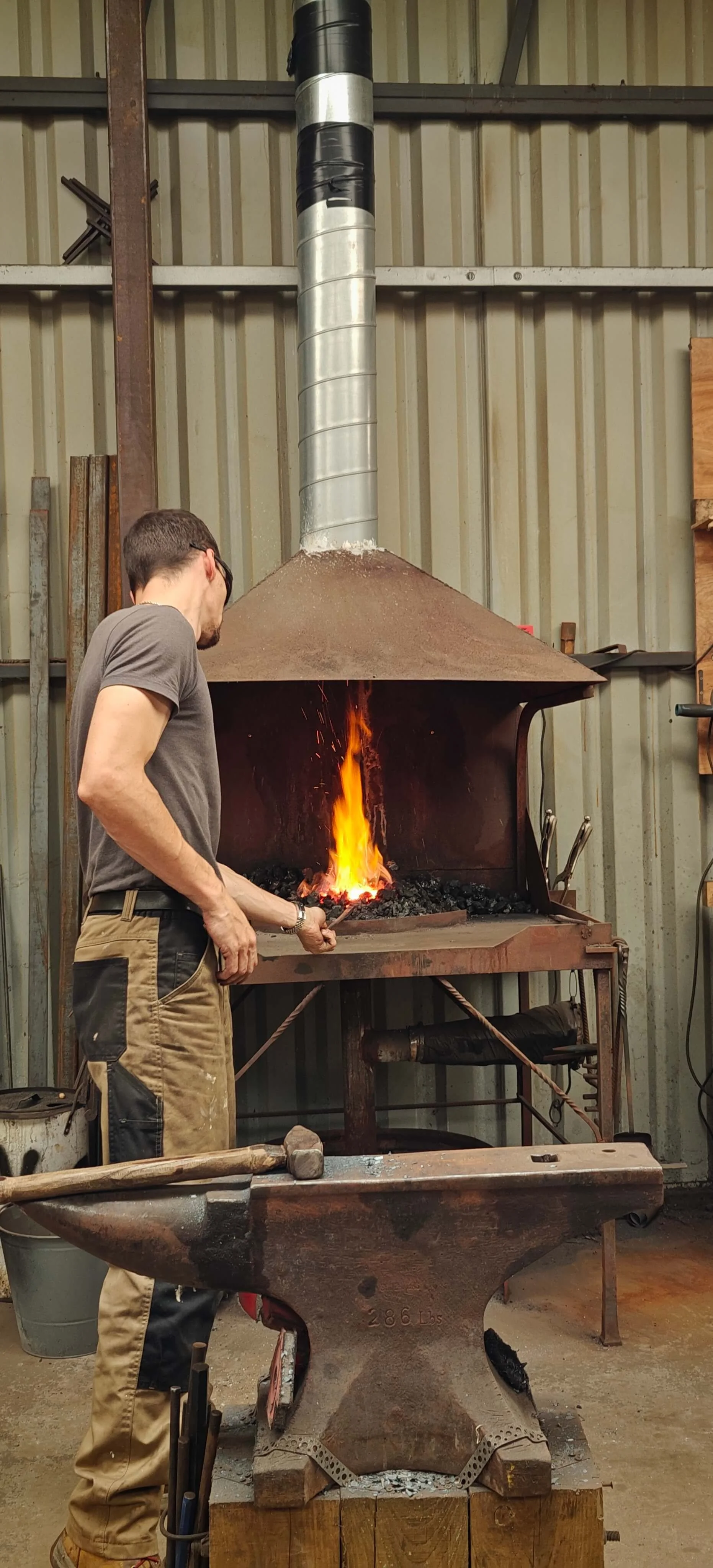 A blacksmith hand-forging metal in Gabha na Lámh Fada (Long Arm Forge), with a fire glowing inside a forge.