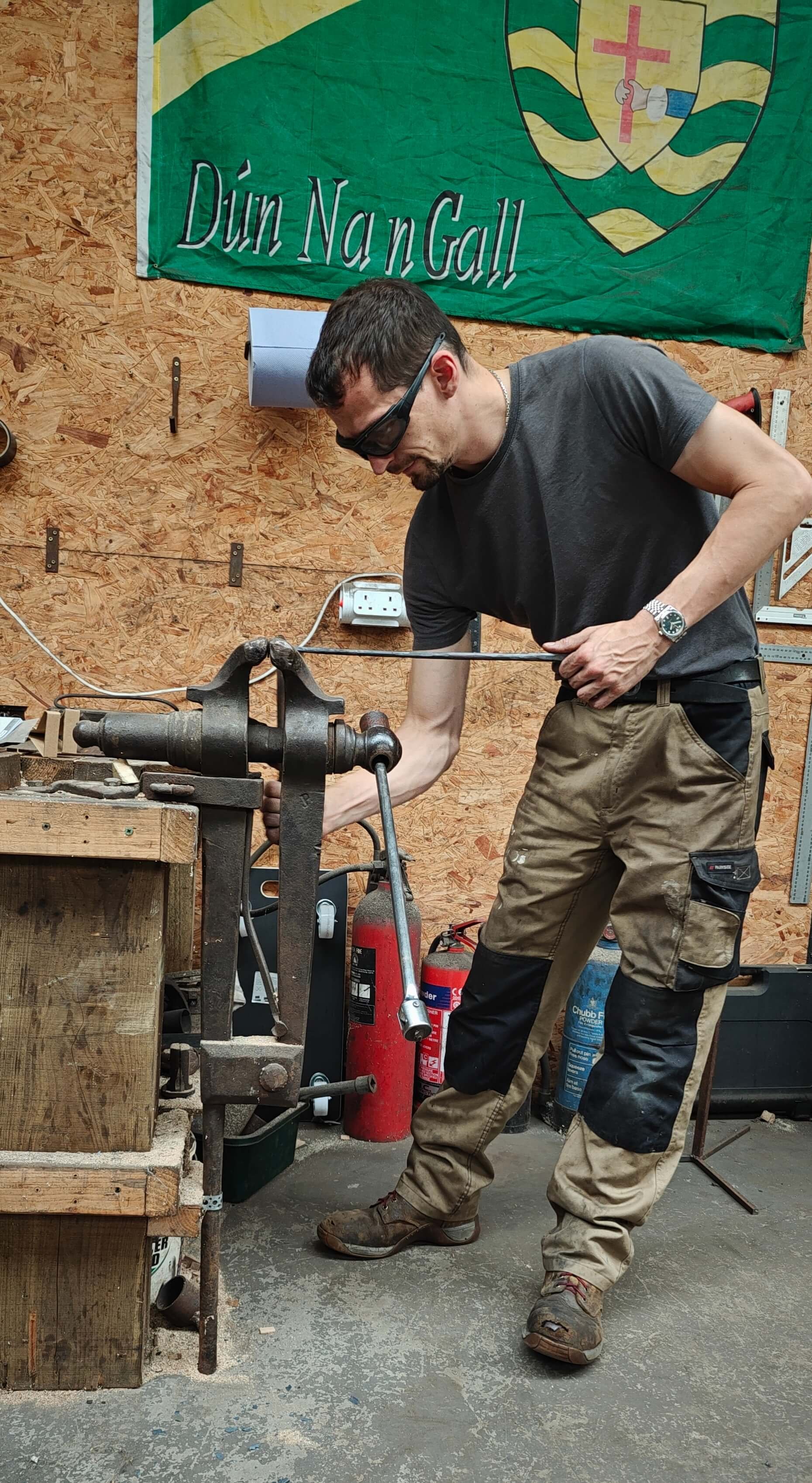 A blacksmith using a metal vice in Gabha na Lámh Fada (Long Arm Forge) while working on a piece of metalwork with various tools and wooden walls.