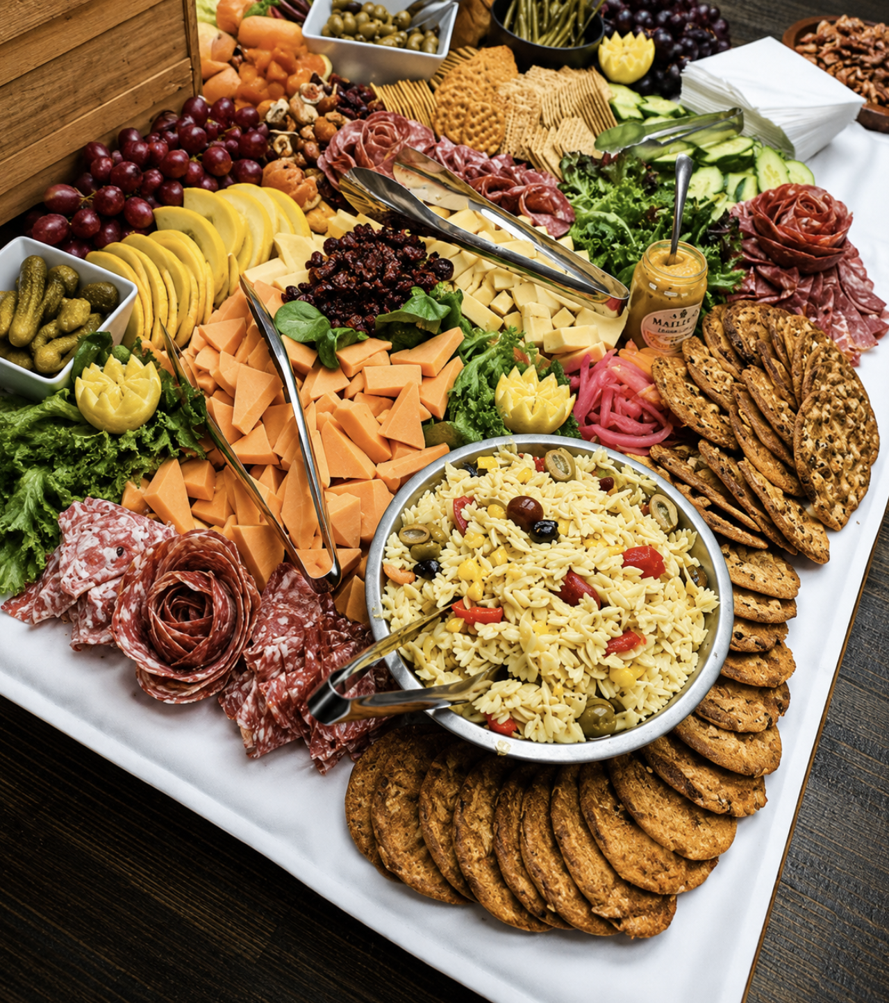 A large platter with an assortment of cheeses, sliced meats, crackers, pasta salad, fresh vegetables, pickles, grapes, and a jar of mustard, arranged for a buffet or party.