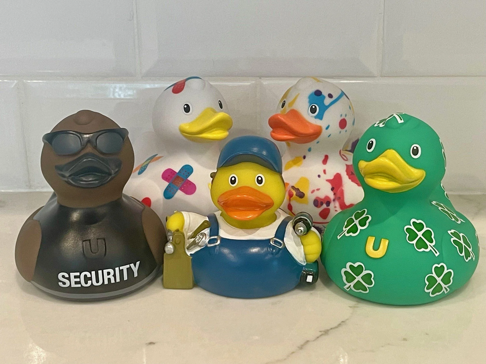 A collection of six rubber ducks with various themes and accessories, arranged on a marble surface in front of a white tile wall.