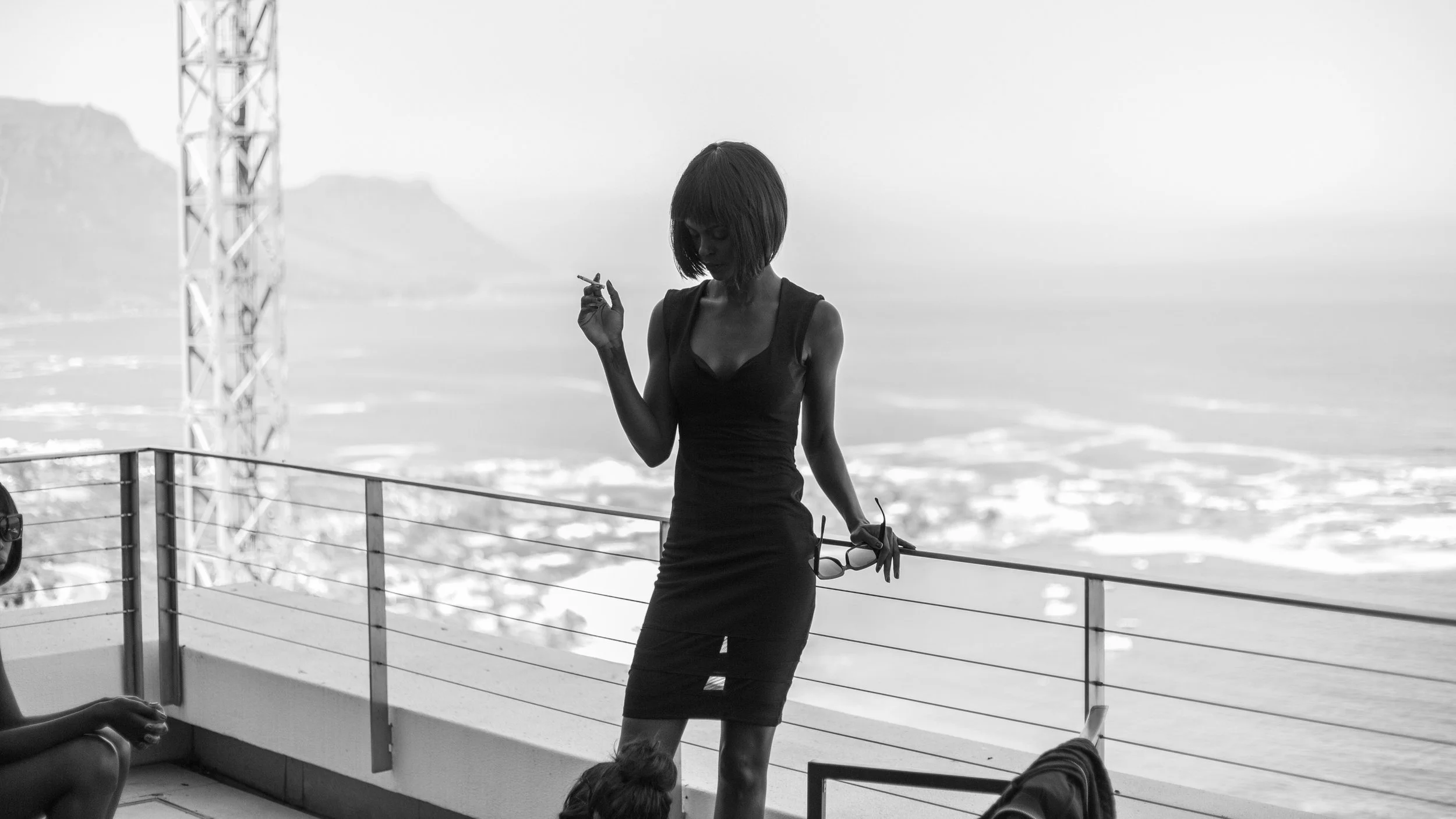 Silhouette of a woman in a black dress holding sunglasses, standing on a balcony with a coastal view; monochrome photo.