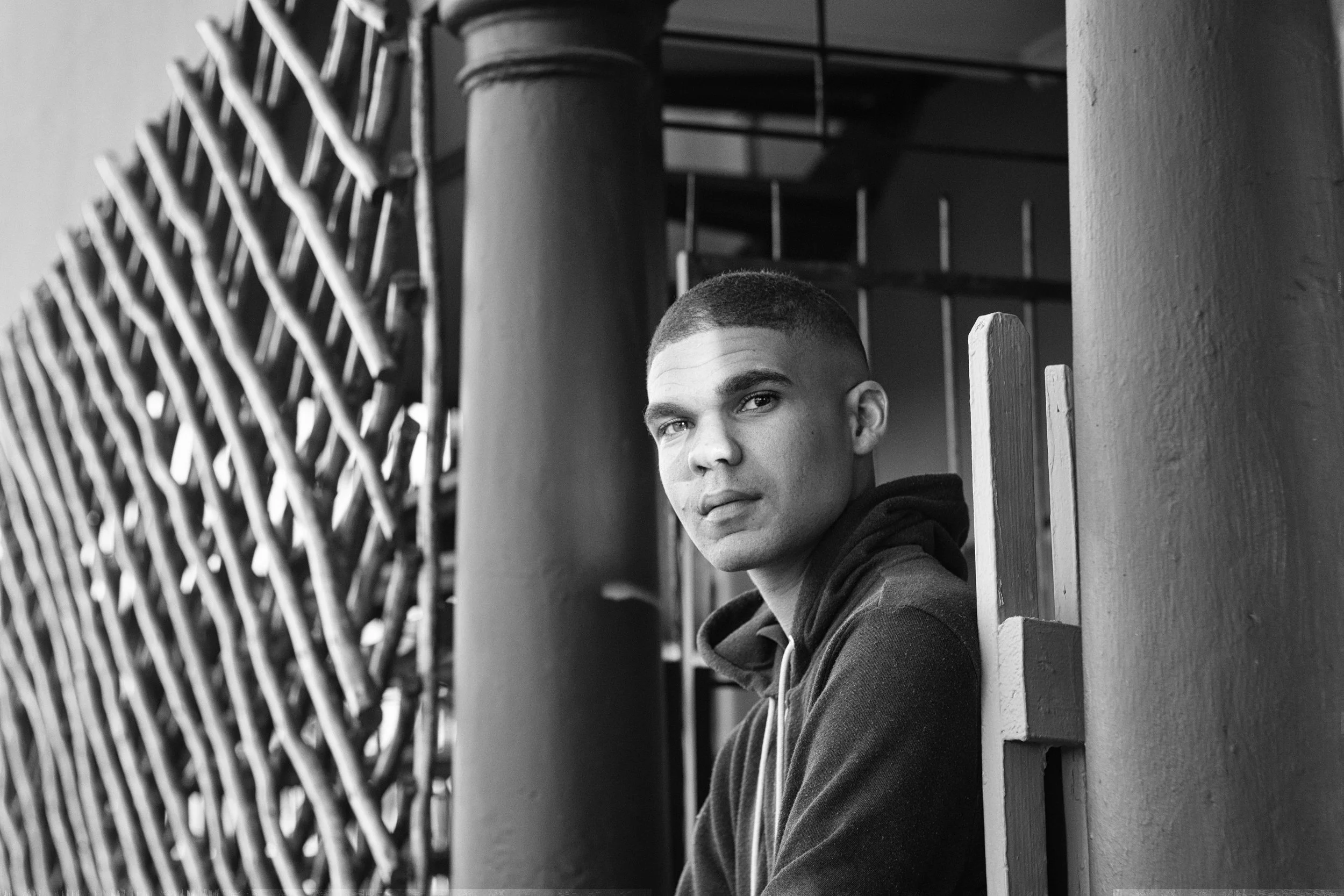 Black and white portrait of a person sitting by a column with a woven fence in the background.