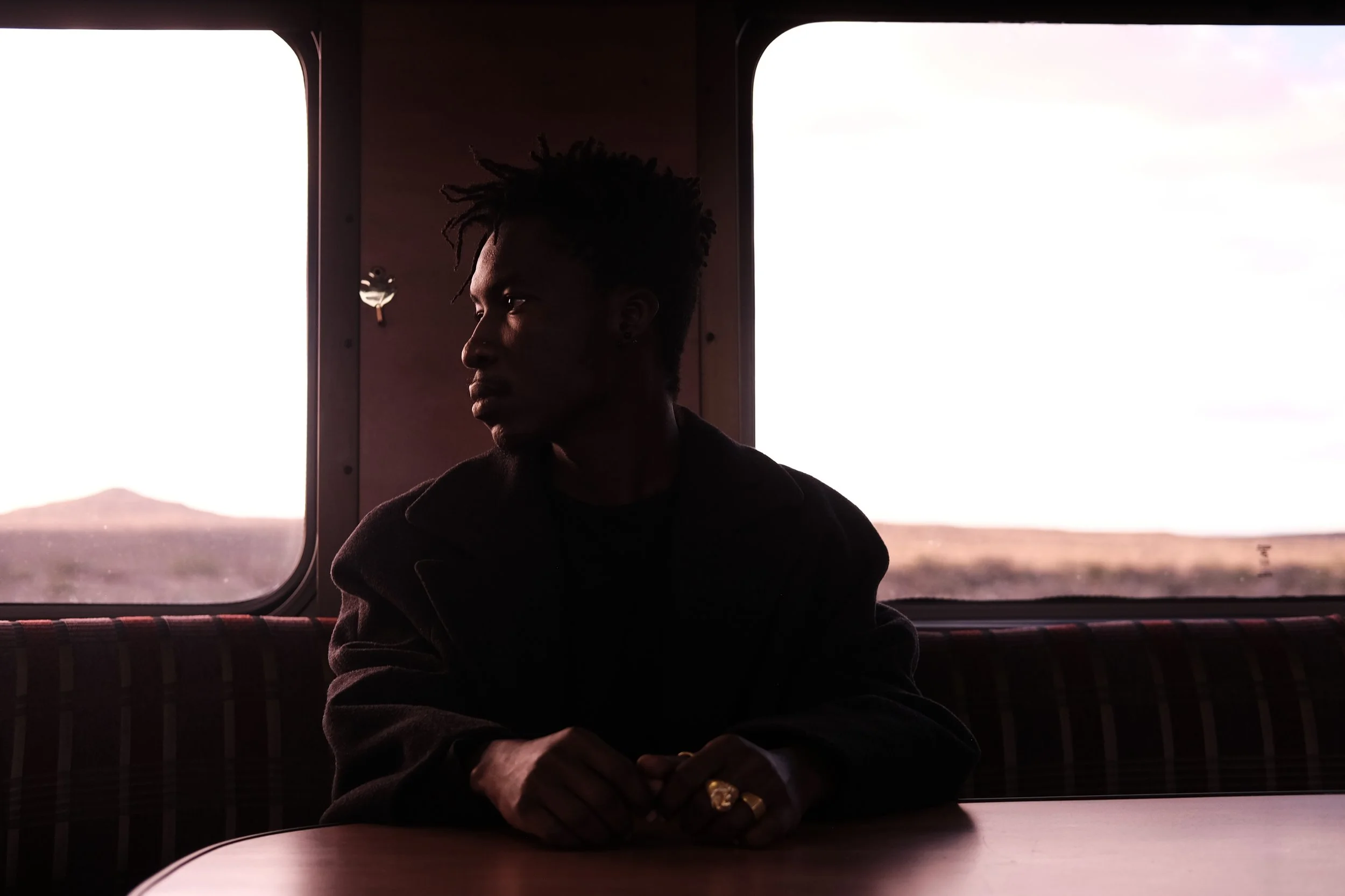 Person sitting in a dimly lit train compartment, looking out the window at a distant landscape during sunset.