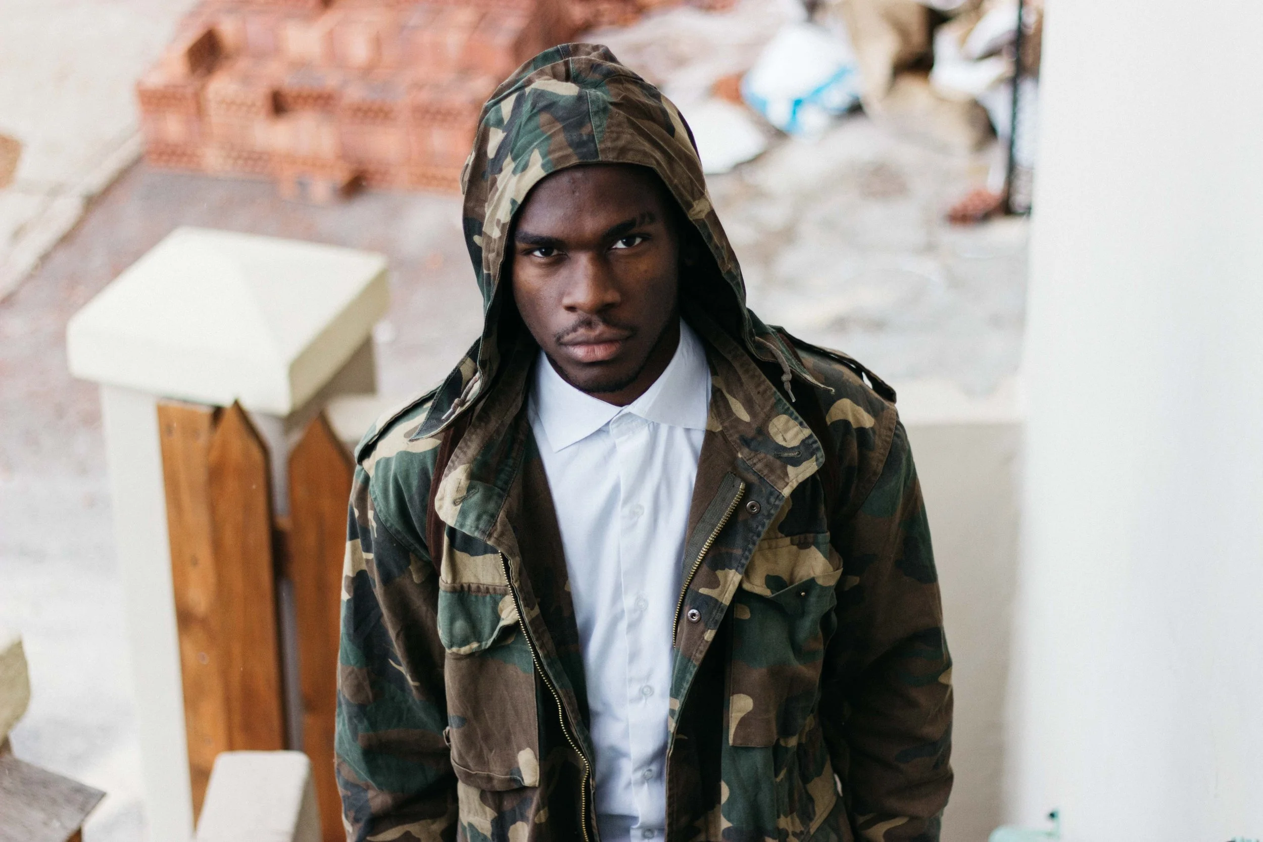 Man in camo jacket and hood standing on staircase with bricks in background