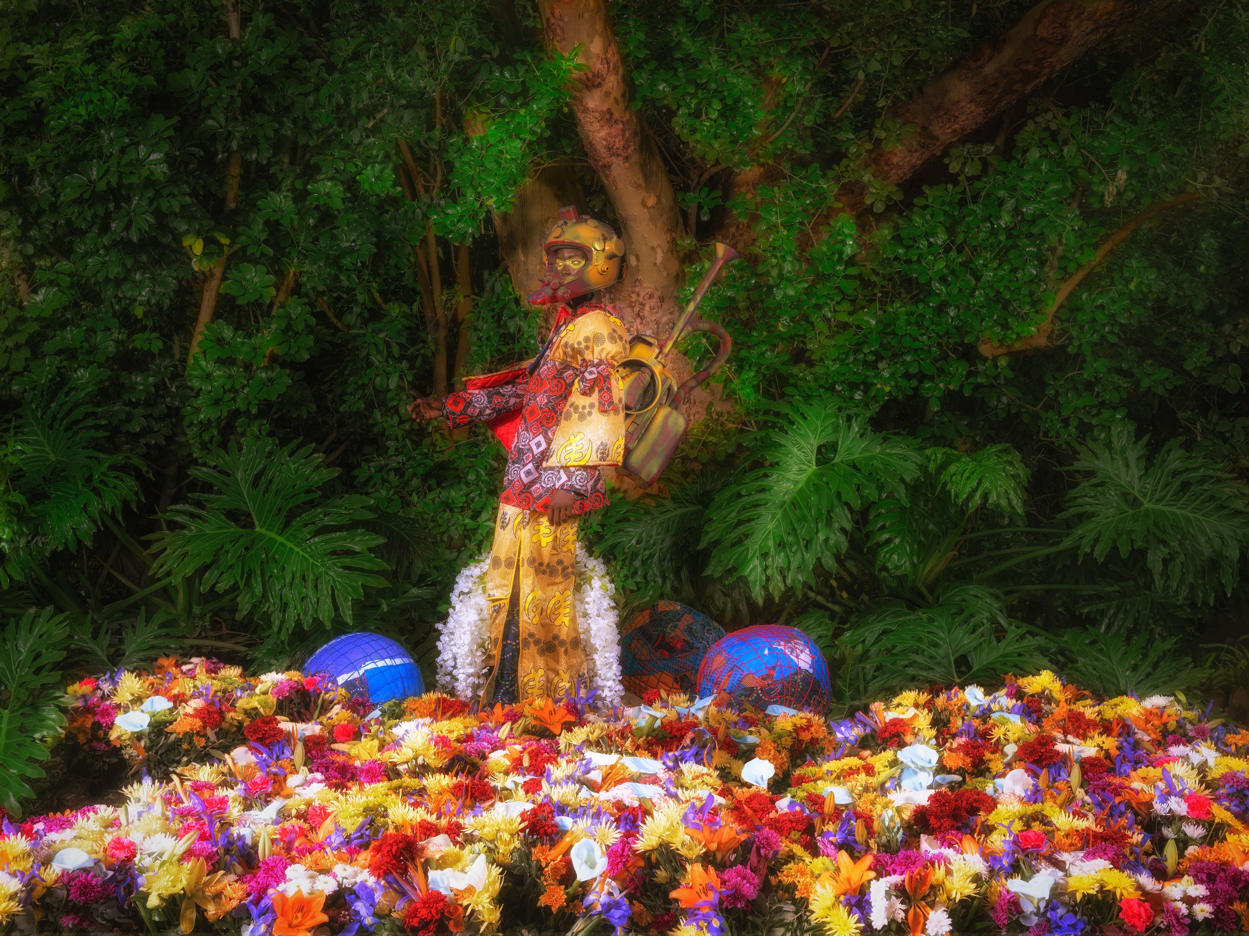 Vibrant art installation featuring a robot-like sculpture with colorful garments and a trumpet, surrounded by an abundance of multicolored flowers and lush greenery.