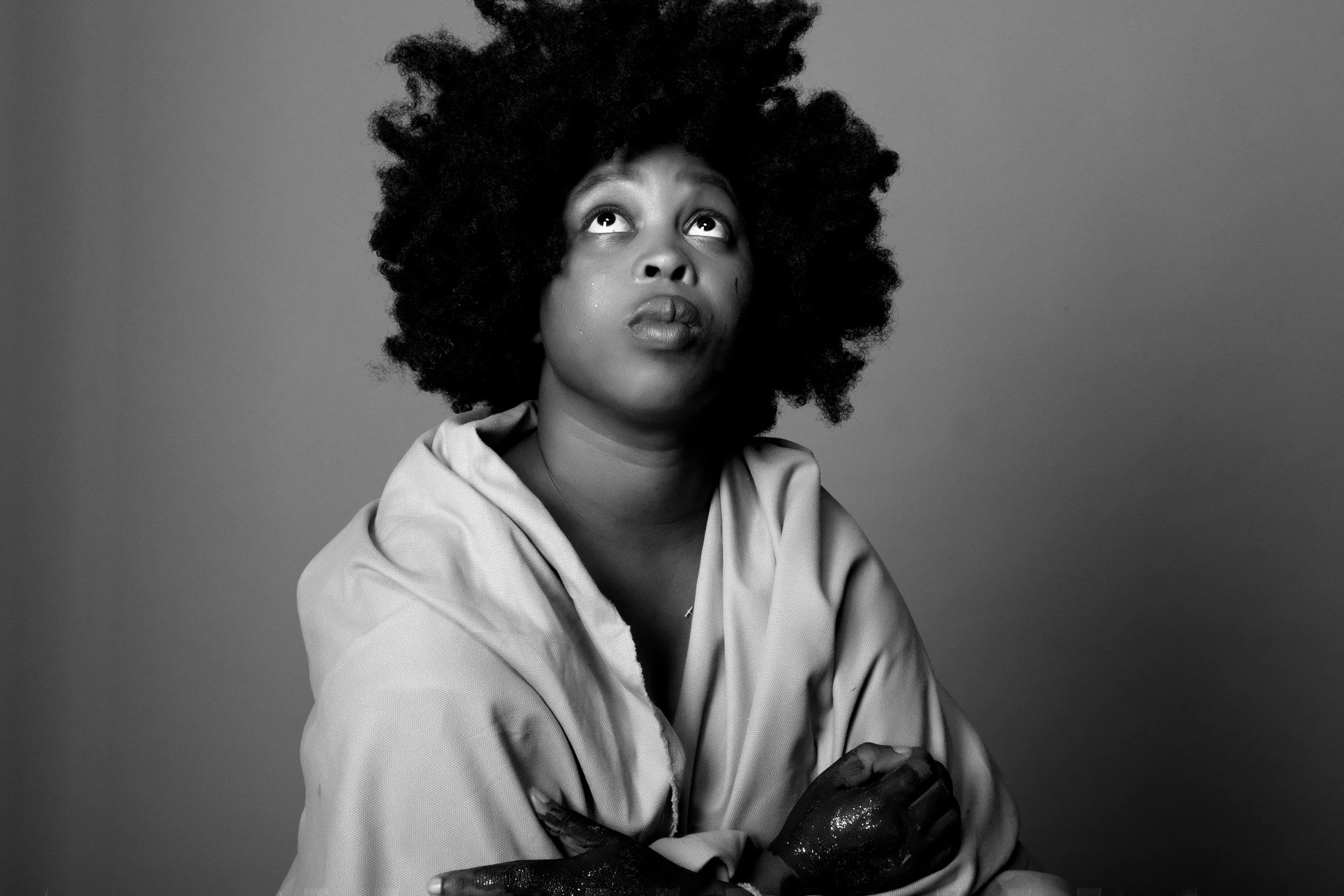 Black and white portrait of a person looking upward, wearing a light-colored garment with arms crossed and hands sparkling.