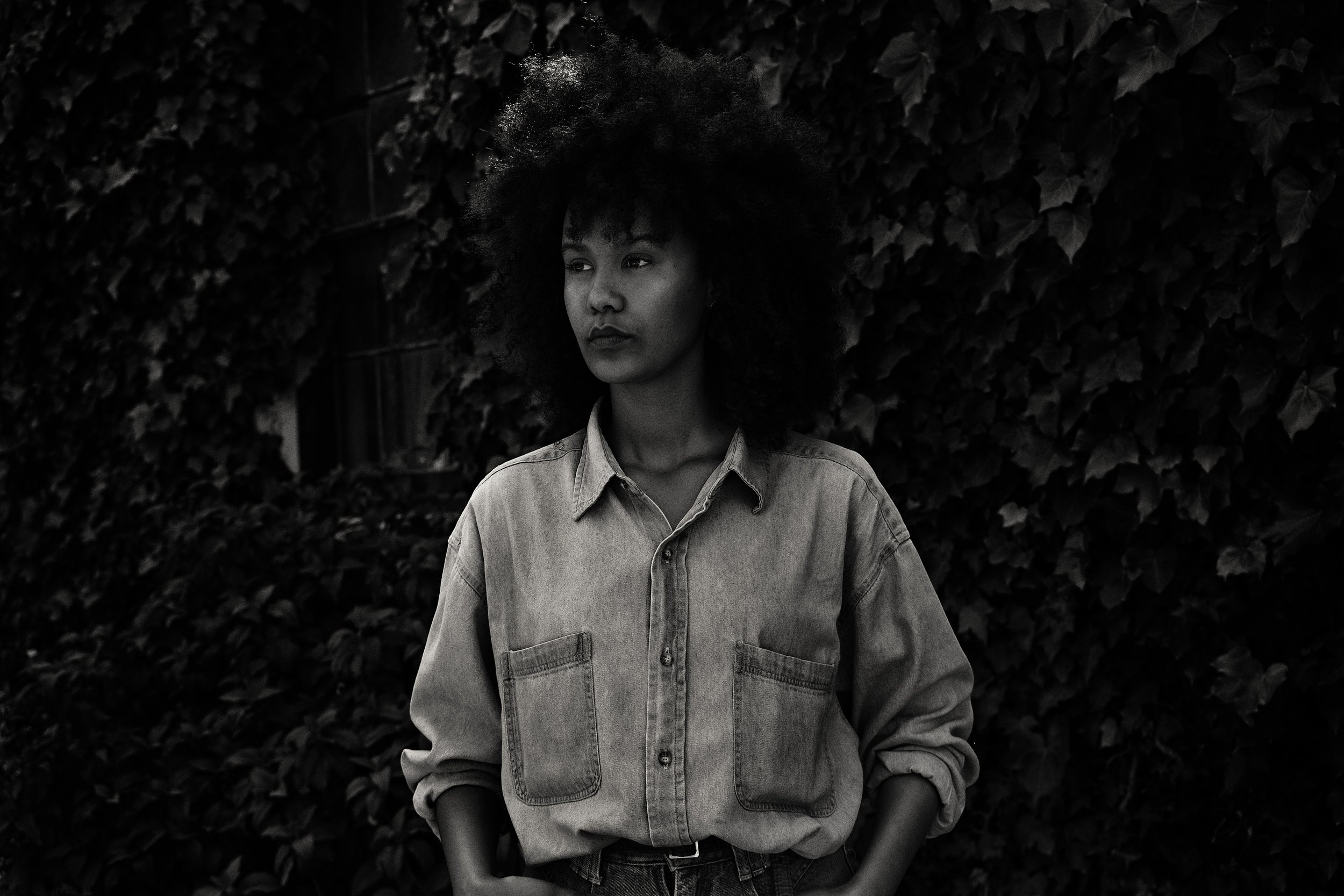 Black and white portrait of a person with curly hair wearing a denim shirt, standing against an ivy-covered wall.