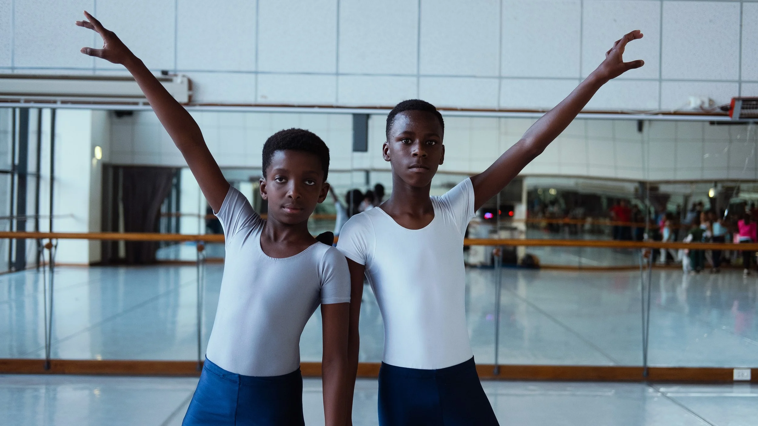Two young male ballet dancers in dance studio, wearing light blue leotards, posing with arms raised, large mirror in background.