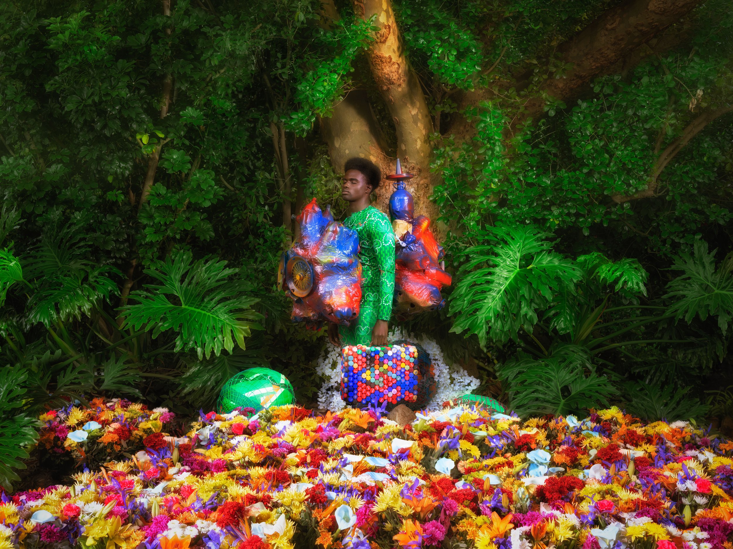 Person standing amid colorful artificial flowers, holding a suitcase made of bottle caps, surrounded by lush greenery.