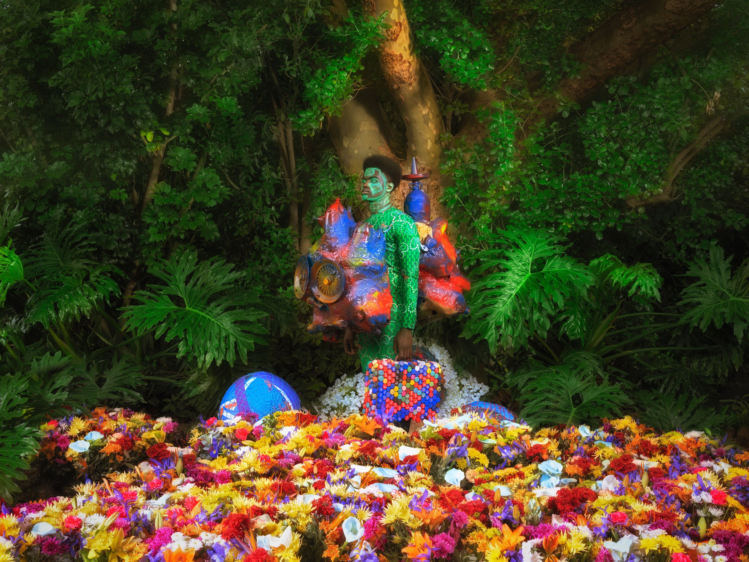 A person with intricate body paint blending into nature, surrounded by colorful flowers and a vibrant abstract structure with large circular elements. Dense green foliage forms the backdrop.