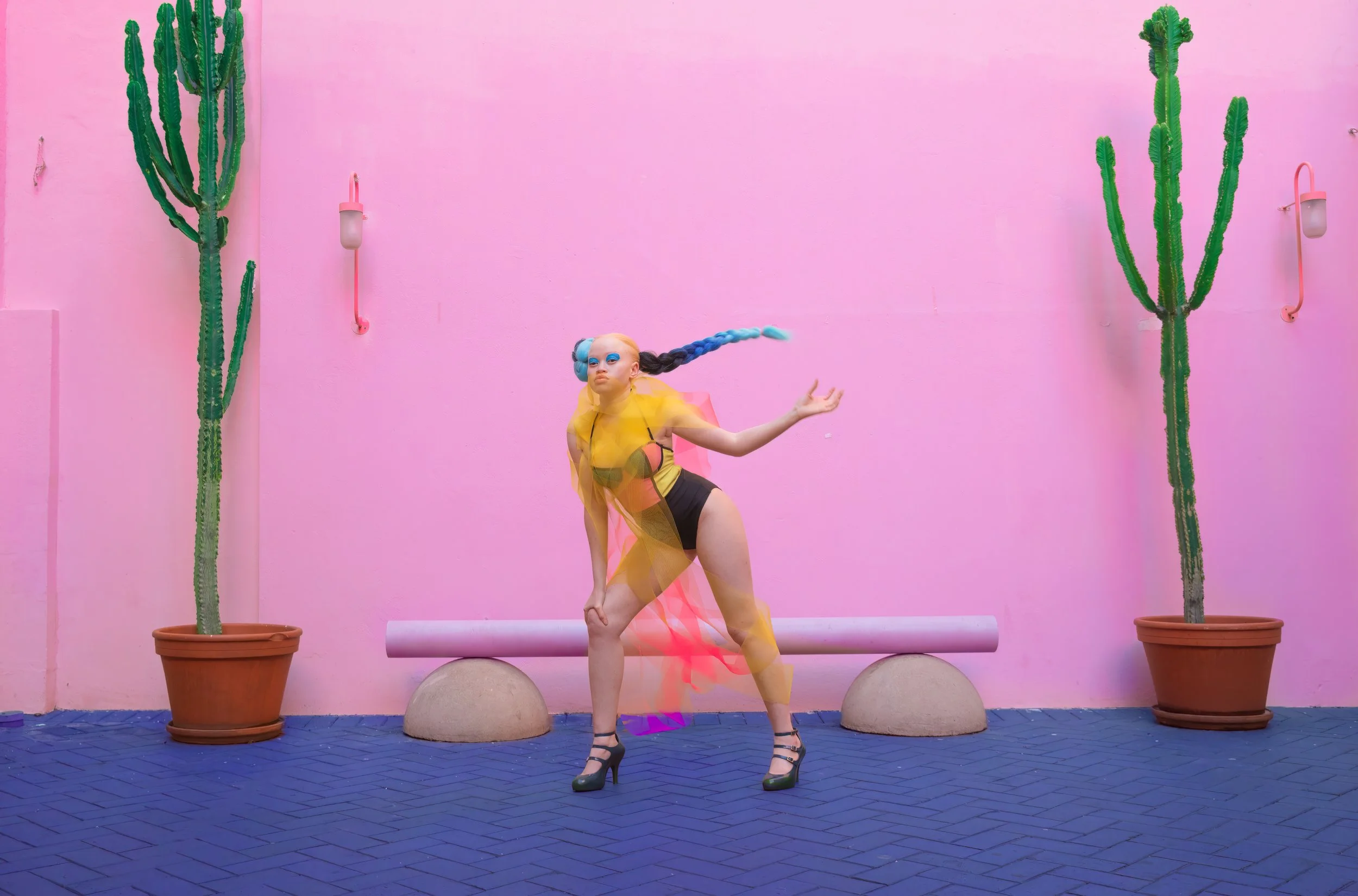Person with colorful braids and vibrant outfit posing in front of pink wall with potted cacti.