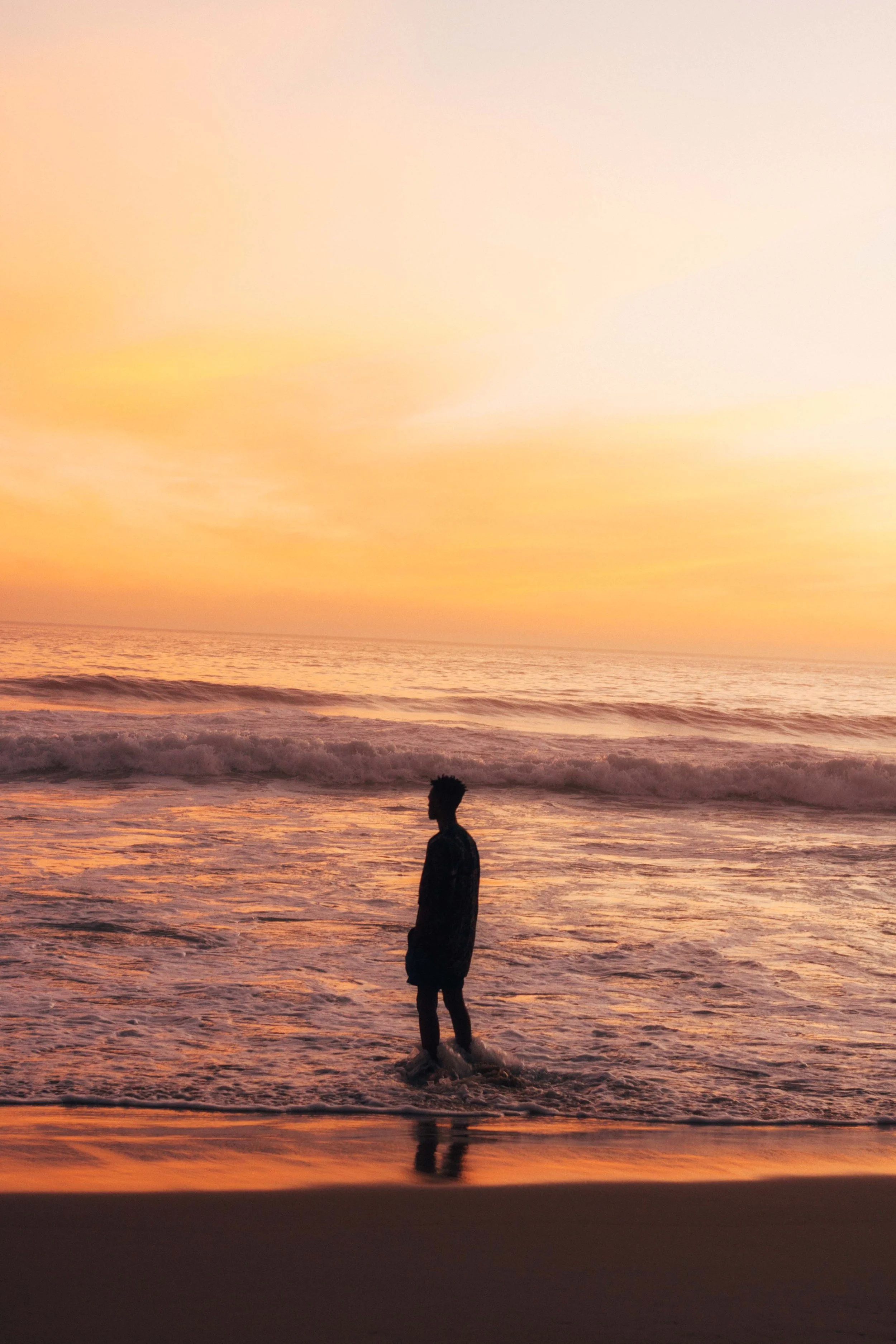 Silhouette of person walking on beach at sunset with waves and orange sky.