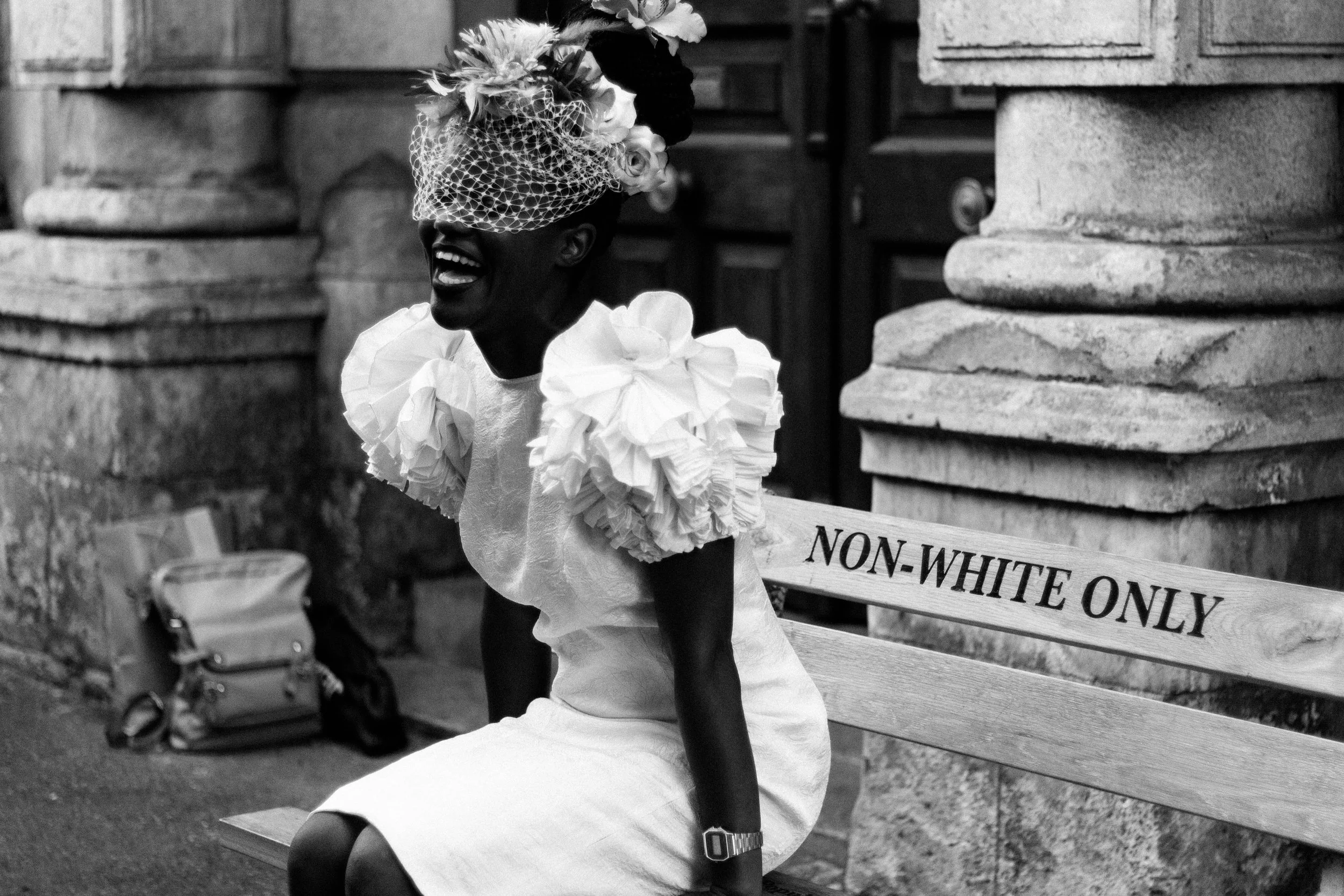 Black and white image of a person sitting on a bench with the words 'Non-White Only.' The person is smiling, wearing an ornate hat and a dress with ruffled sleeves.