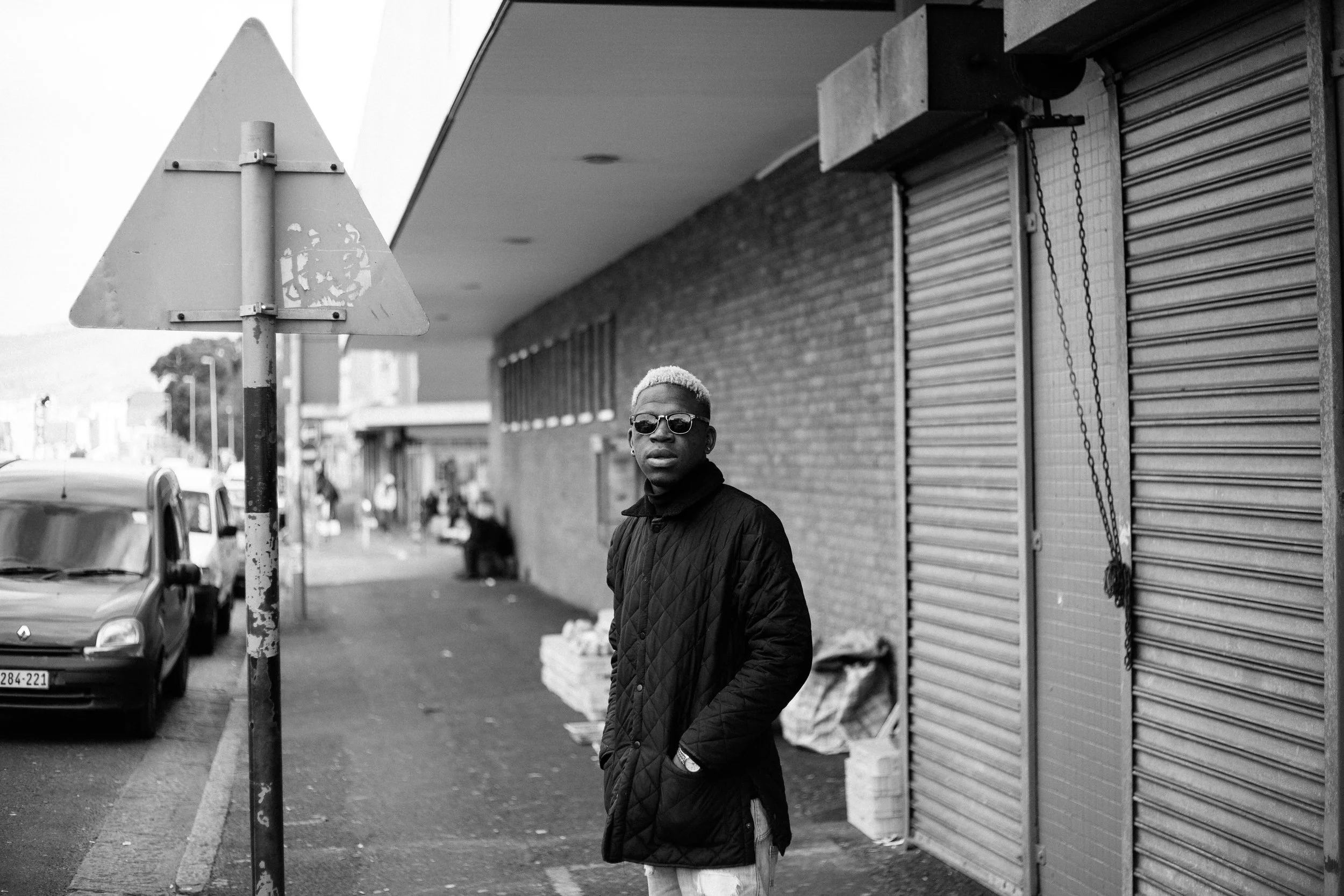 Black and white street scene with person wearing sunglasses and coat, standing near closed shop shutters.