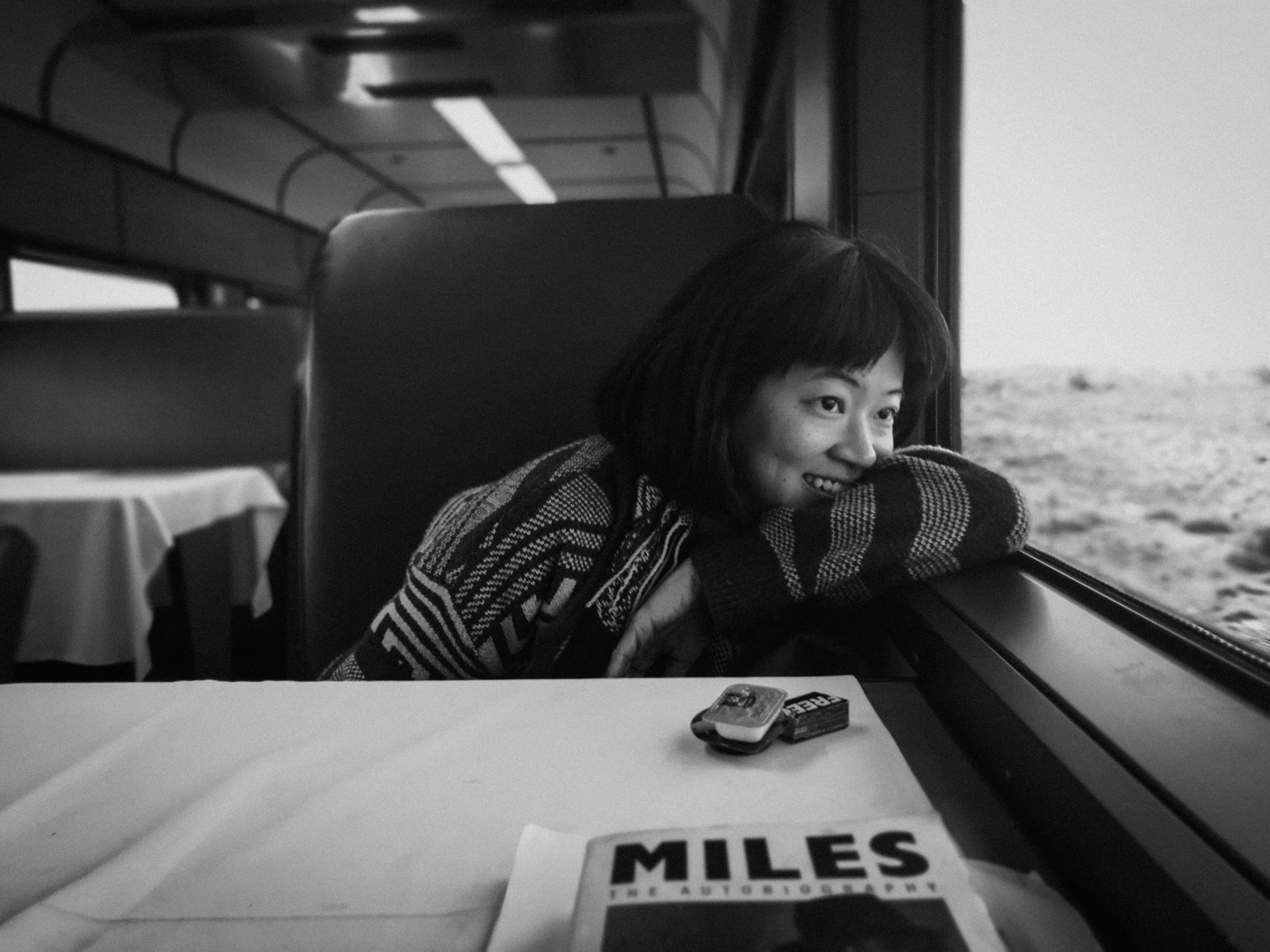 Black and white image of a person smiling, looking out a train window, with a book titled "Miles" and small items on the table.