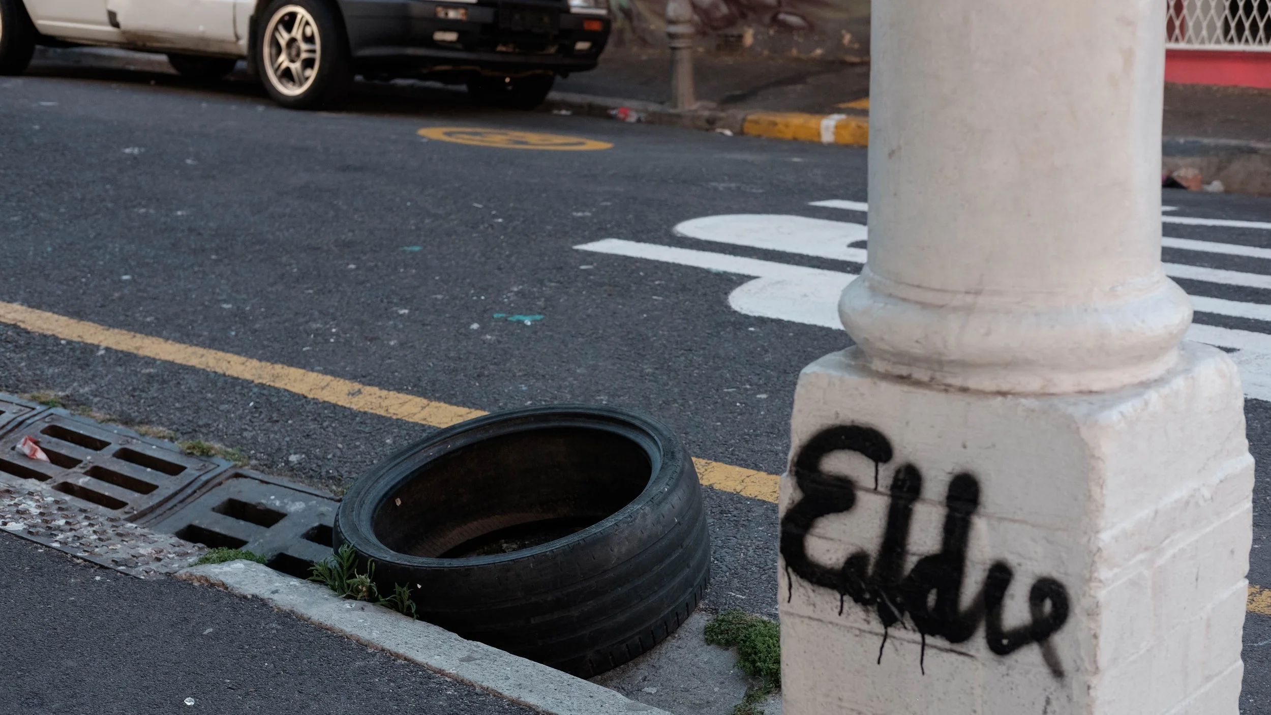 Street scene with white pillar, graffiti, discarded tire, parked car, and road markings.