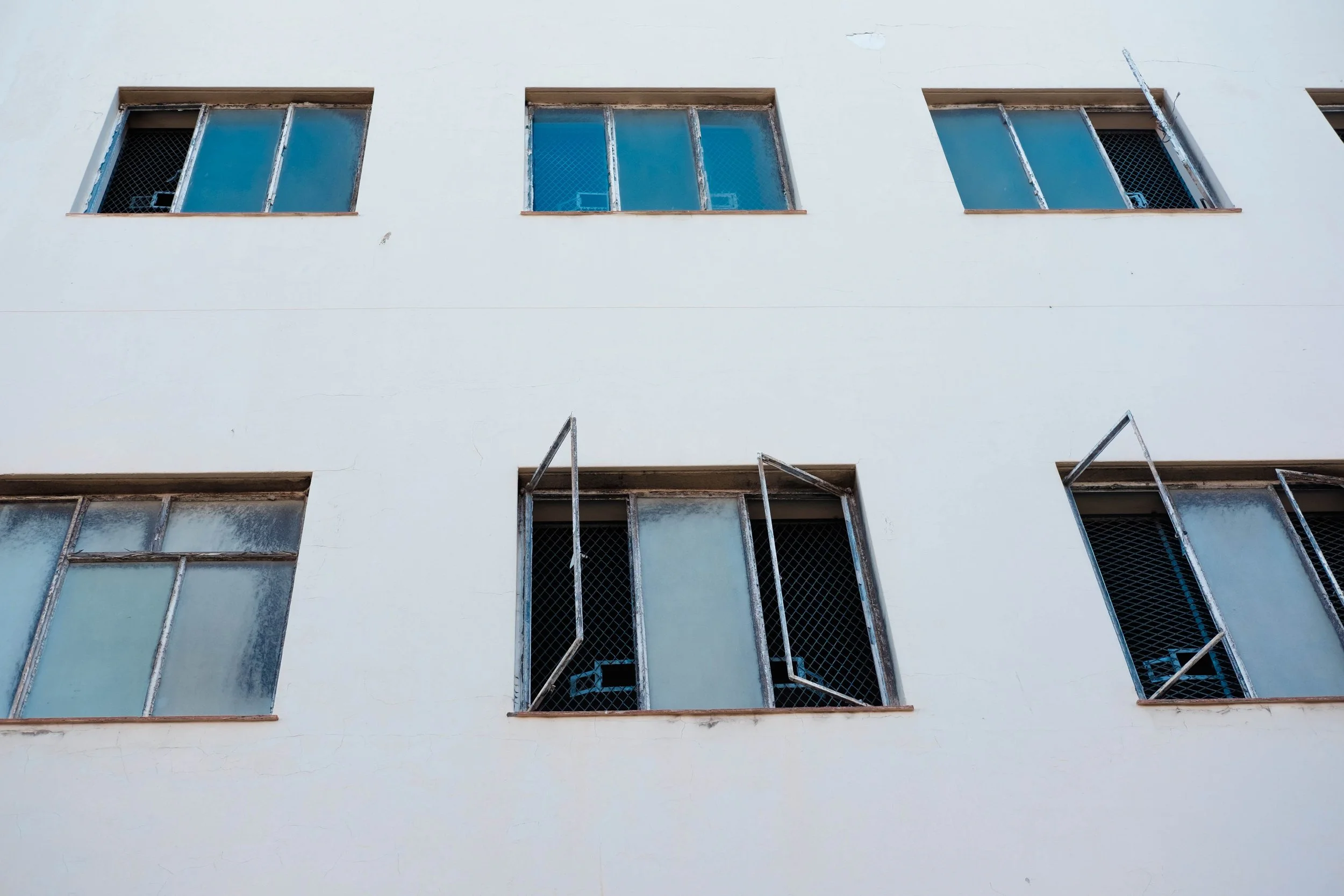 Building facade with six windows, some open, with metal grills and blue-tinted glass.