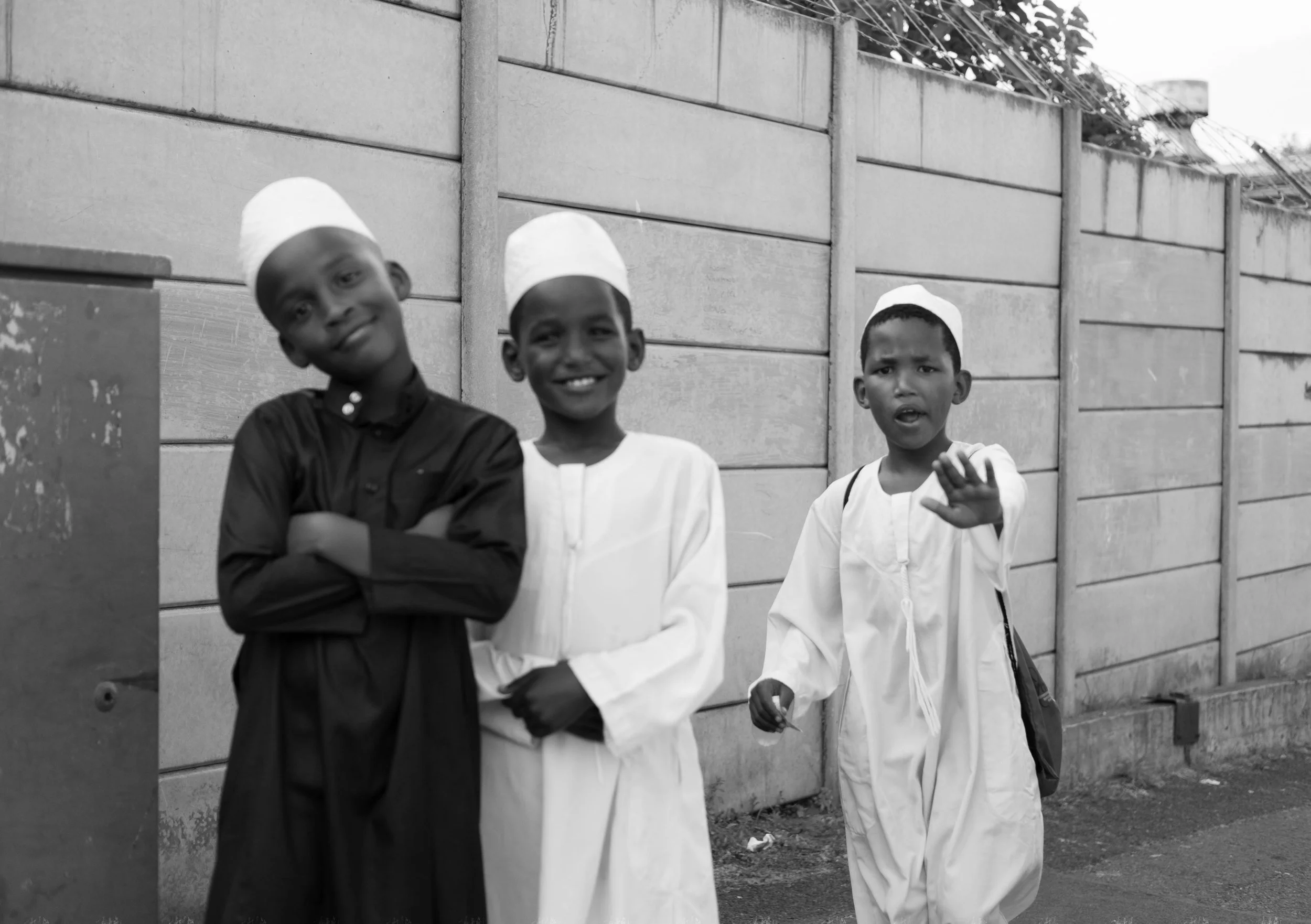 Three boys in traditional clothing walking along a wall street in black and white.