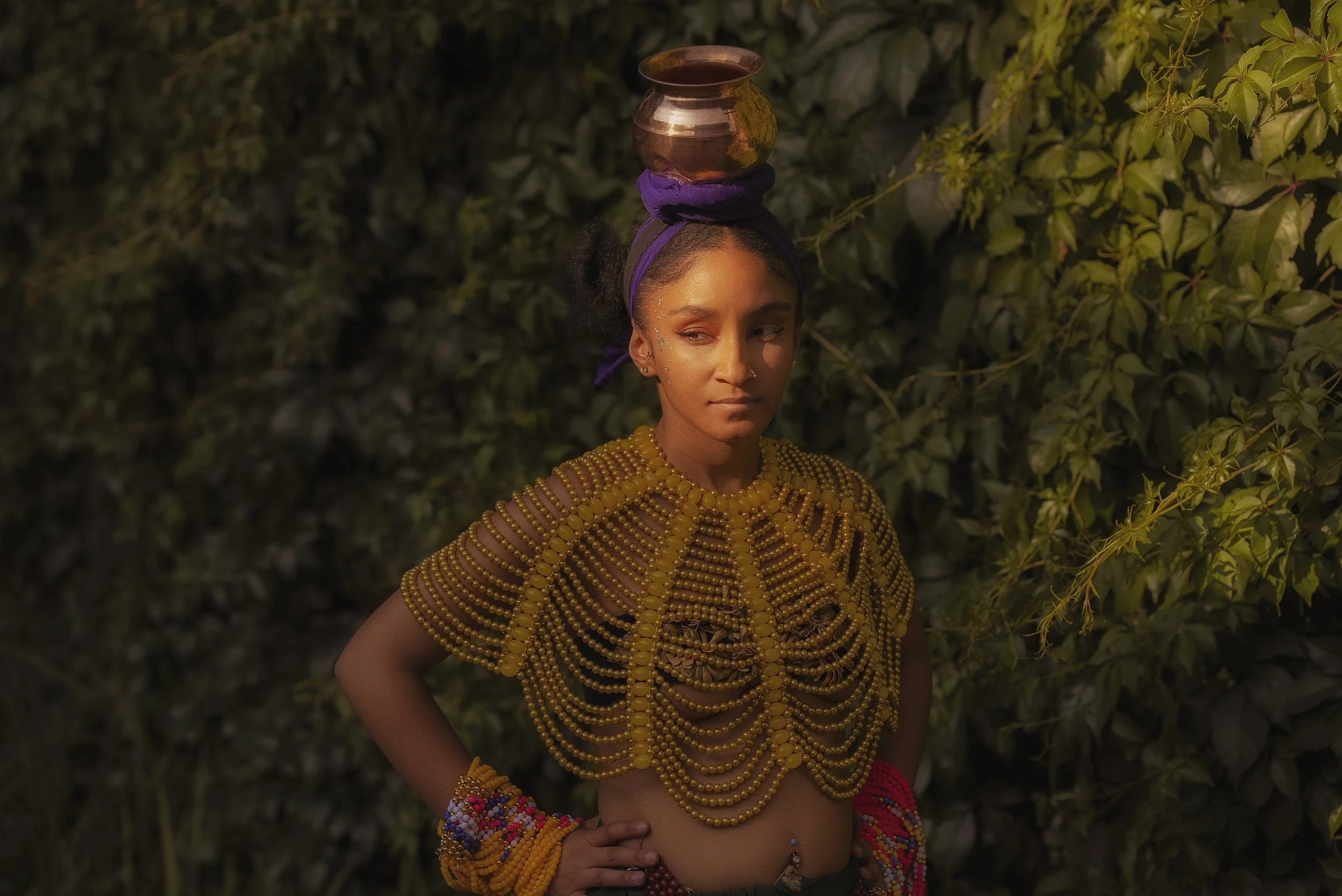 Woman wearing yellow beaded top with copper pot on head, standing against leafy background.
