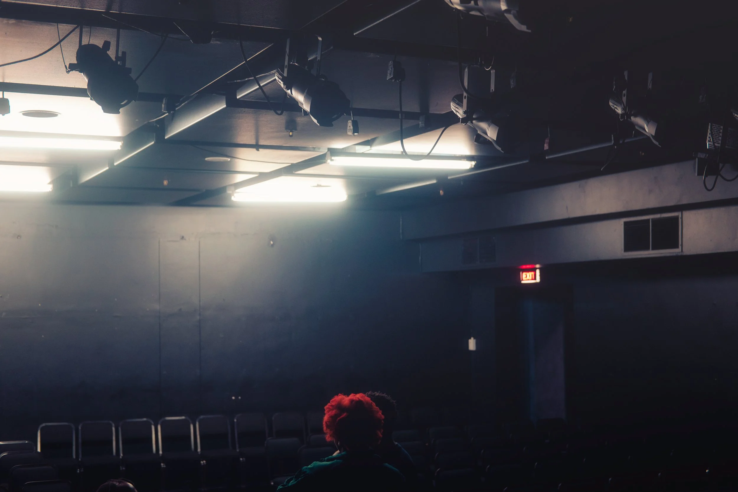 empty theater with stage lights, black walls, rows of empty chairs, exit sign