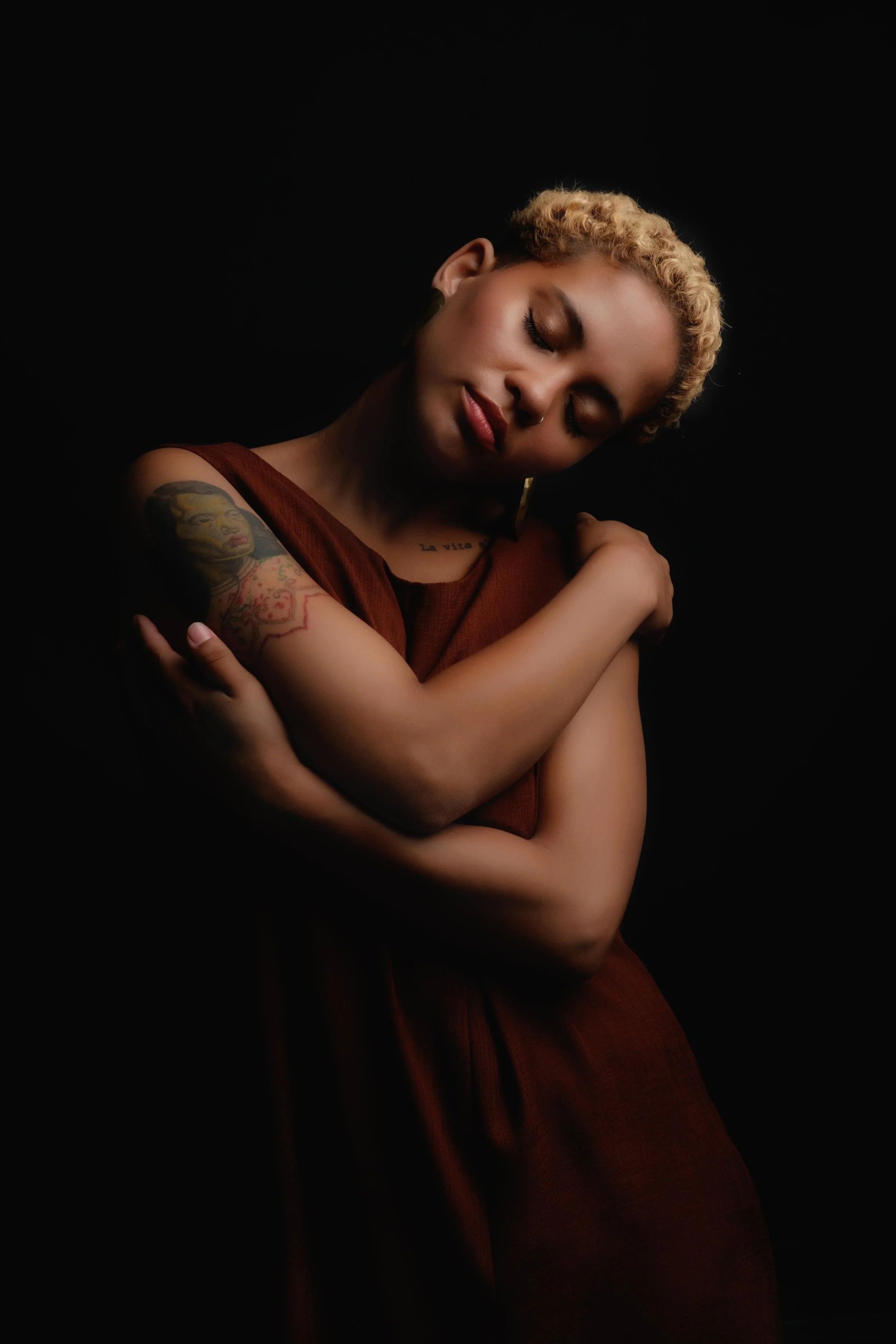 Woman with short curly hair hugging herself, wearing a brown dress, with tattoos on her arm, set against a black background.