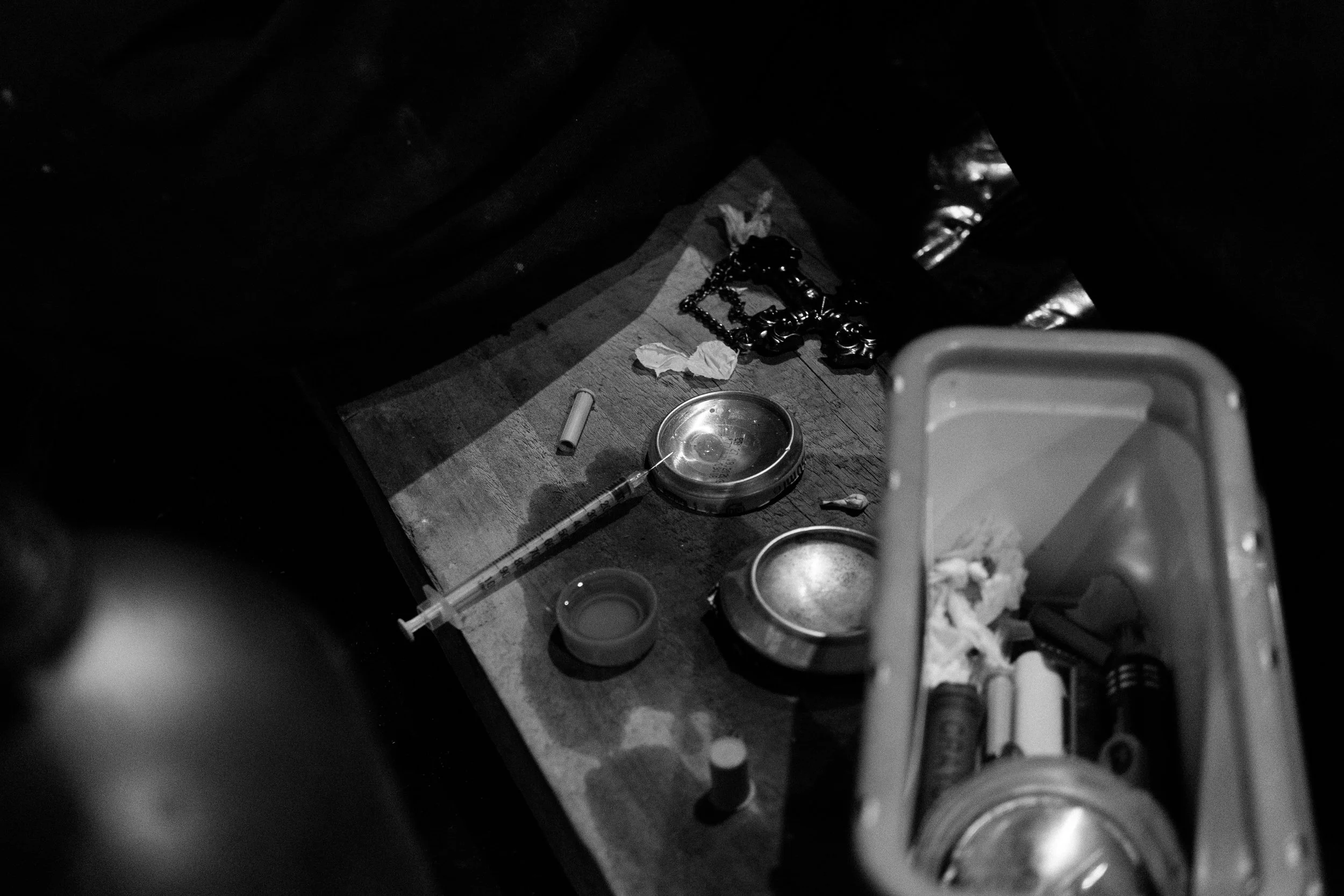 Black and white image of drug paraphernalia, including a syringe, metal containers, and trash bin with miscellaneous items.