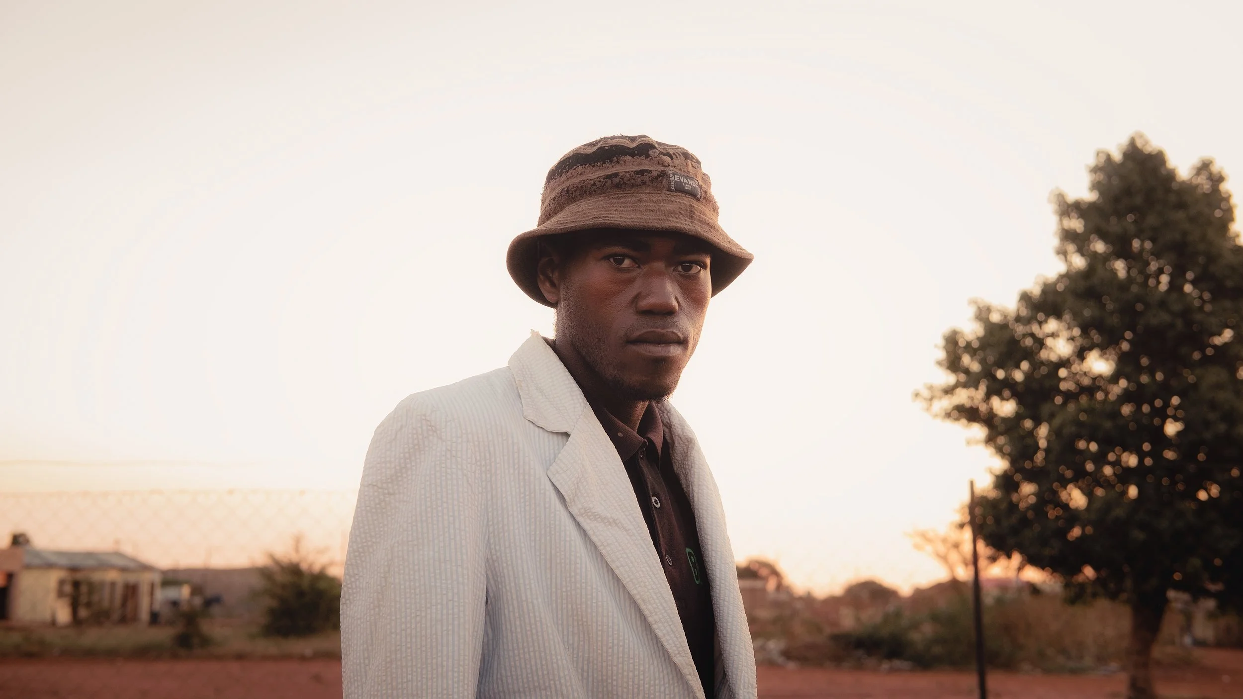 Man wearing a bucket hat and white jacket, standing outdoors with a blurred tree and building in the background during sunset.