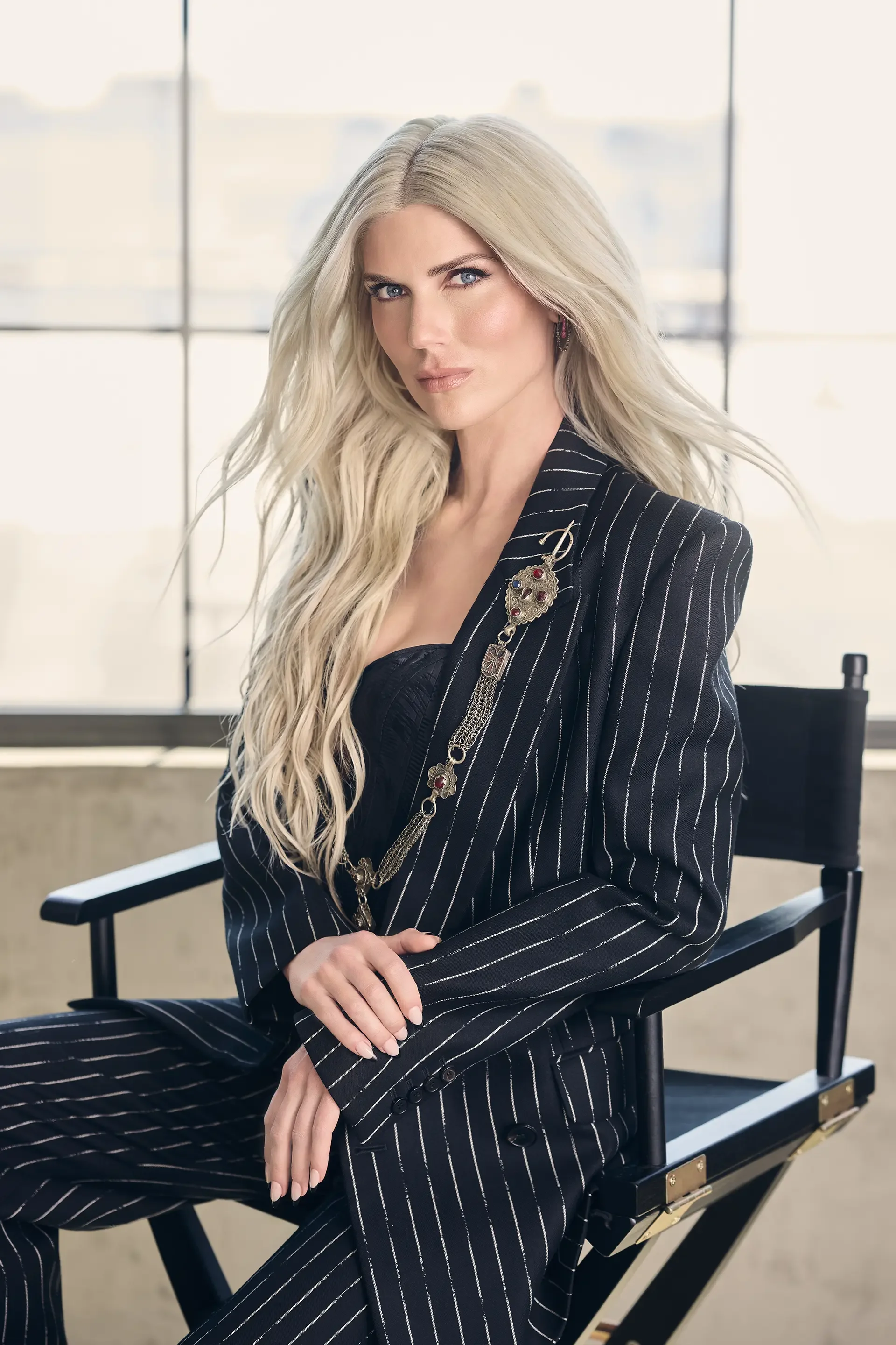 A woman with long, wavy blonde hair and blue eyes, wearing a black pinstripe suit with a decorative brooch on her lapel, sitting on a black director's chair in front of large windows in a bright, modern space.