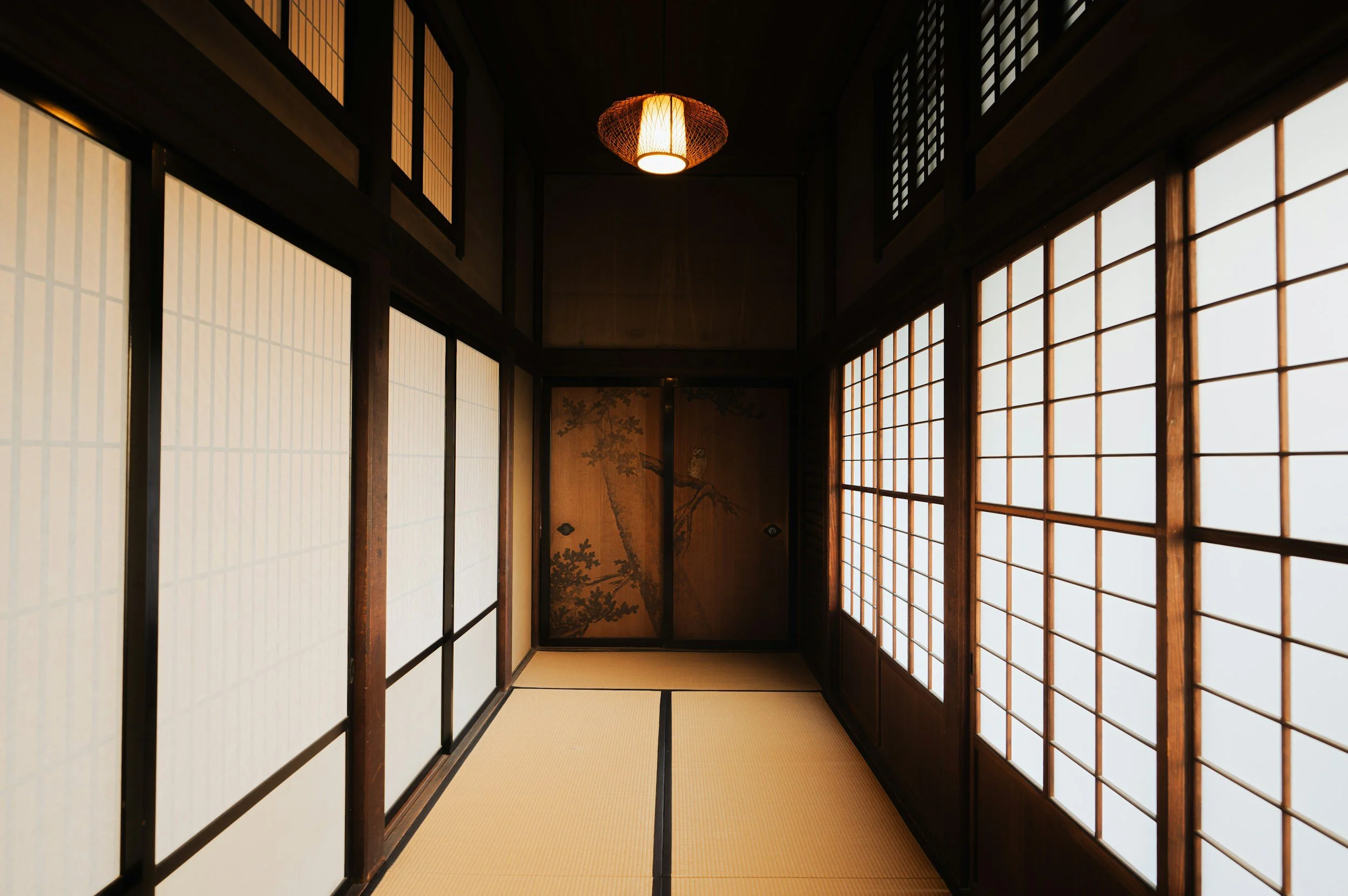 the image looks down a hallway with tatami flooring. On both sides of the image are dark brown shoji doors. At the end of the hall is a pair of painted wooden sliding doors.