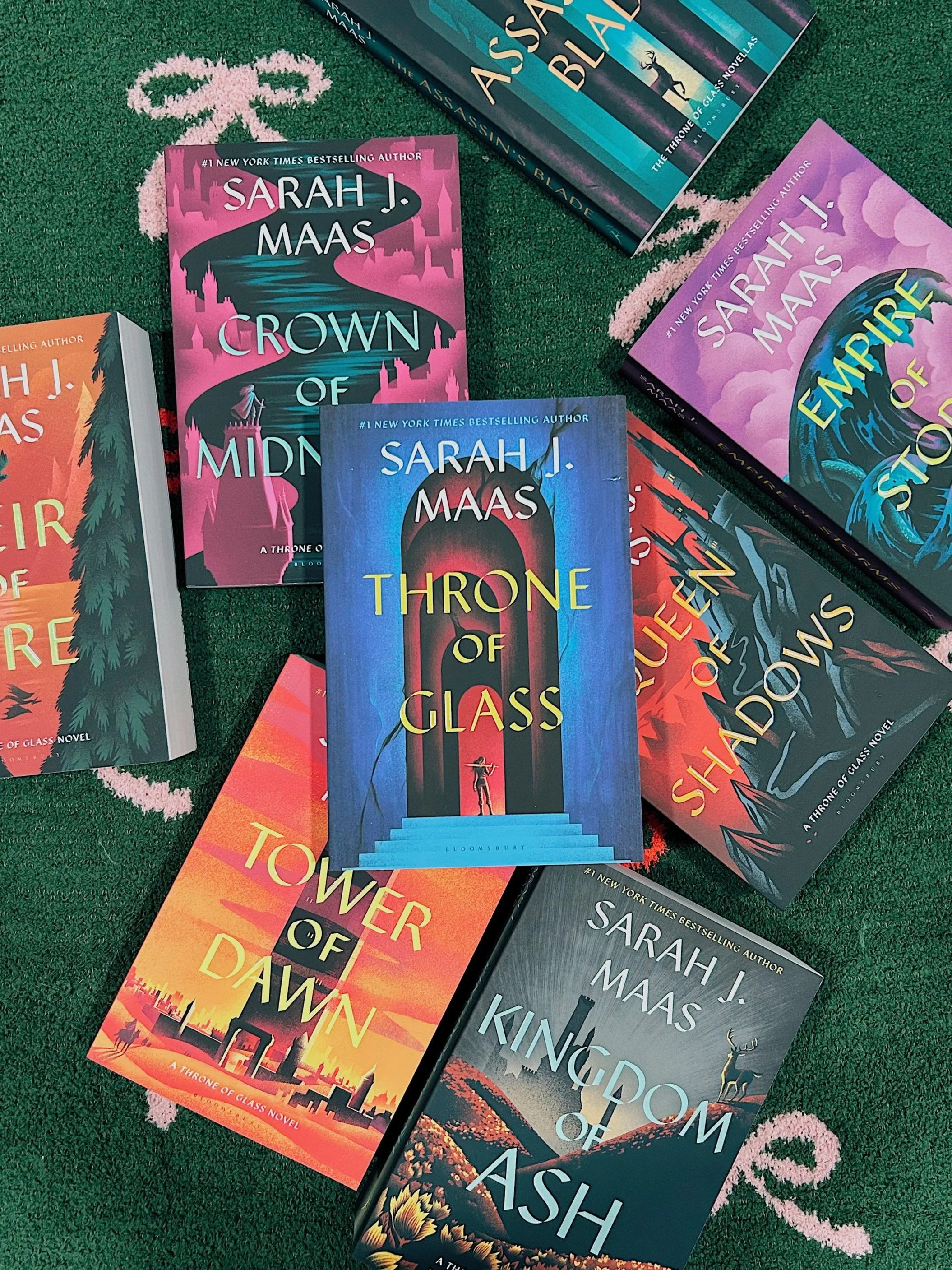 A collection of six Sarah J. Maas books spread out on a green blanket, covered with pink and red bows as the background. The books include 'Crown of Midnight,' 'Throne of Glass,' 'Empire of Storms,' 'Tower of Dawn,' and 'Kingdom of Ash.'