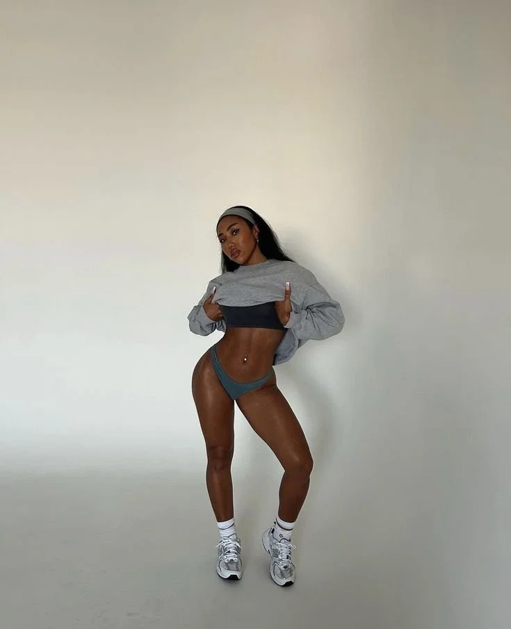 Young woman in athletic wear posing against a plain white background, wearing a cropped sweatshirt, shorts, white sneakers, and a black headband.