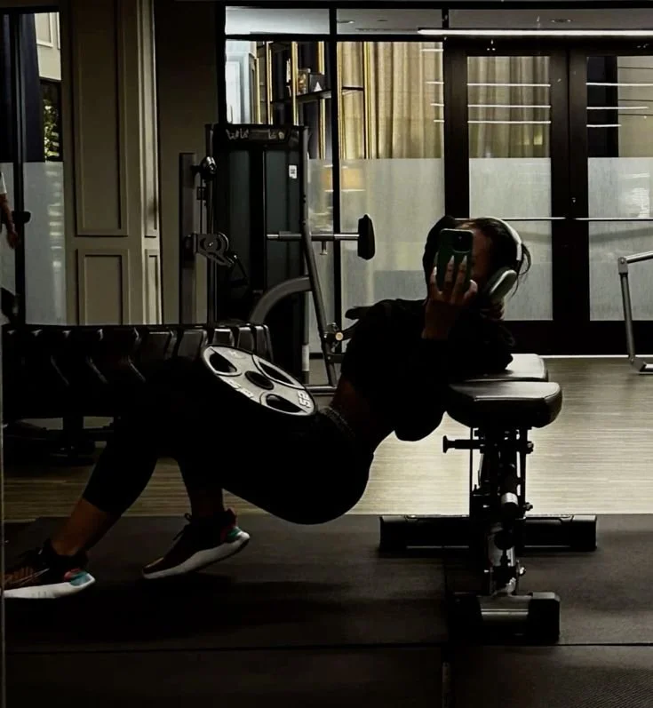 A person taking a mirror selfie at the gym, lying on a workout bench with headphones on, holding a phone to cover their face.