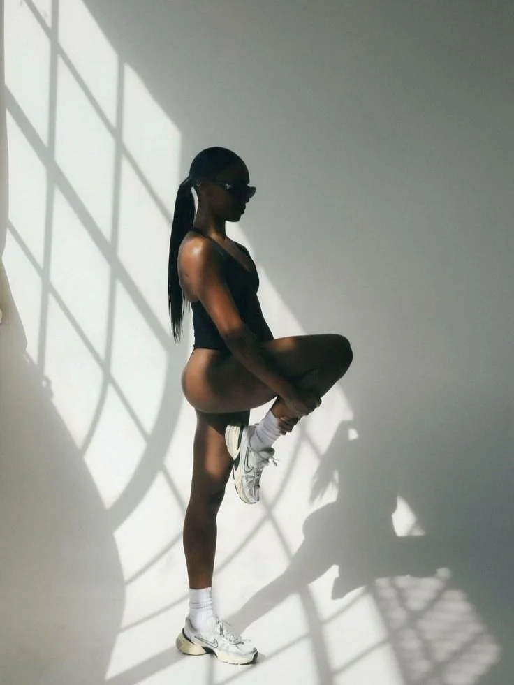 A woman wearing sunglasses, a black athletic top and white sneakers, standing against a white wall with shadows from a grid pattern, lifting her right knee in front of her and balancing on her left leg.