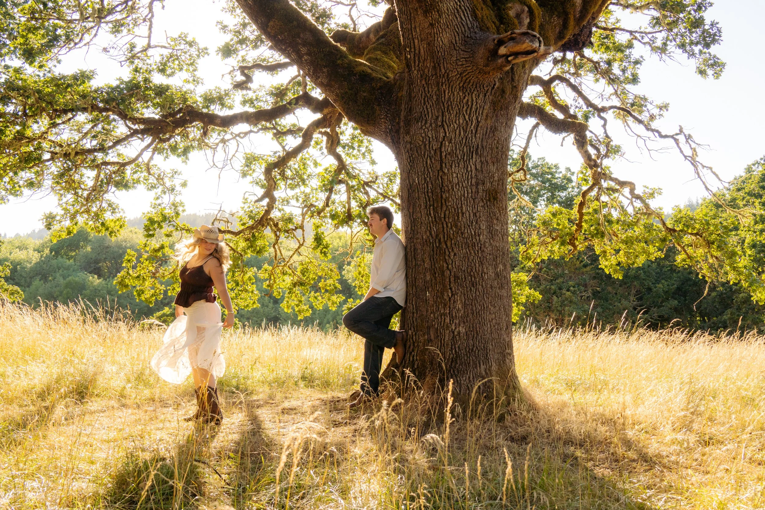 A woman wearing a sun hat and a woman in white clothes standing near a large tree in a field, with another person leaning against the tree, under bright sunlight.
