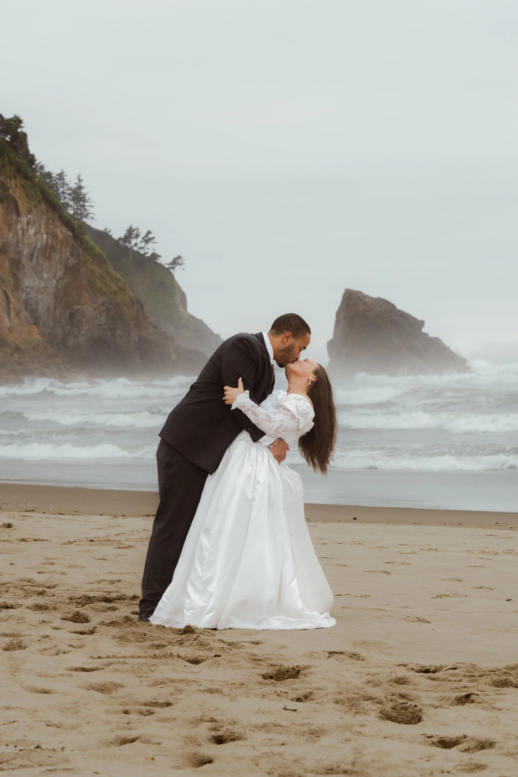 A couple in wedding attire sharing a kiss on Neskowin beach with cliffs and large rocks in the background.