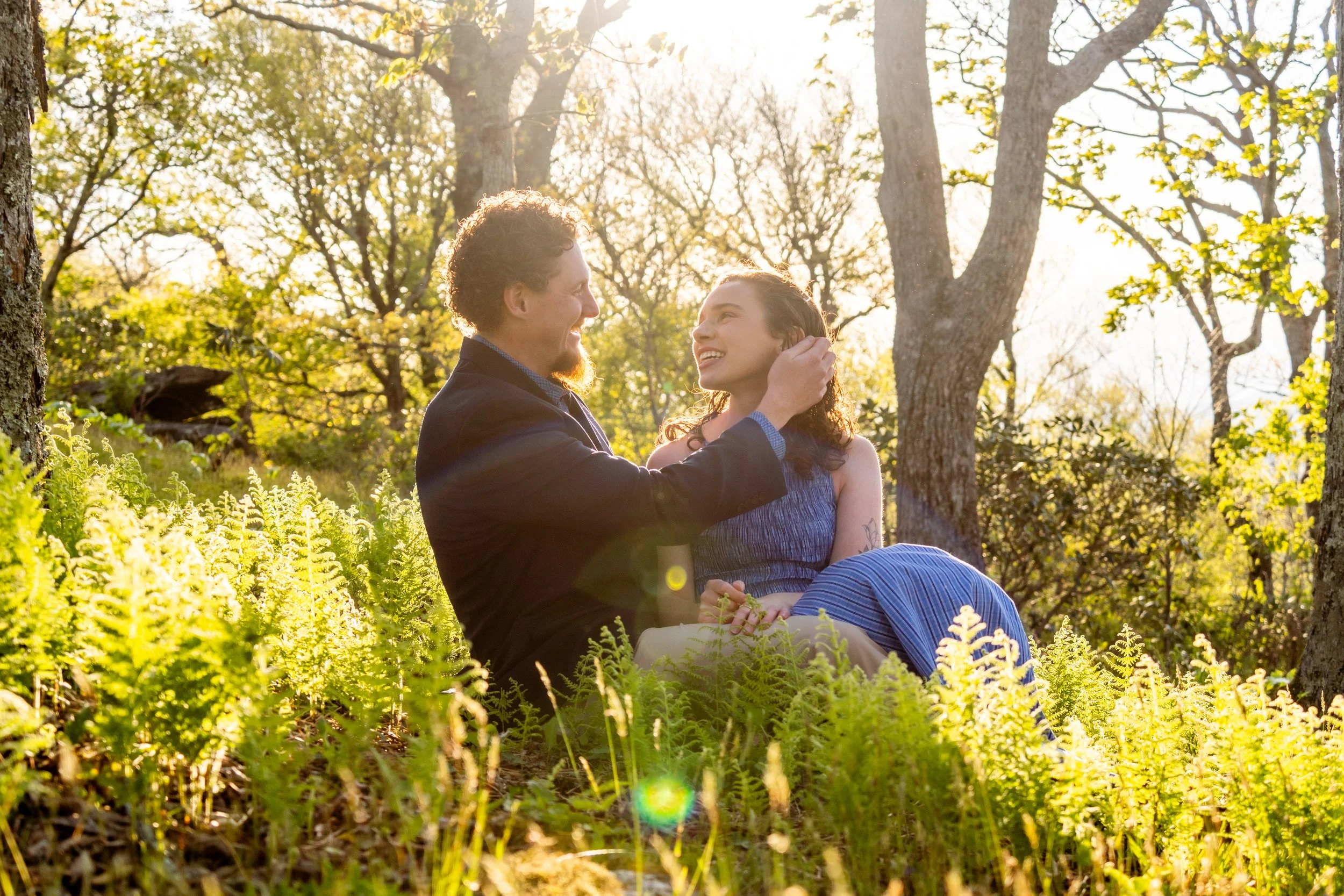 A man and woman sitting on the grass in a forest, smiling and looking at each other, with sunlight filtering through trees in the background.