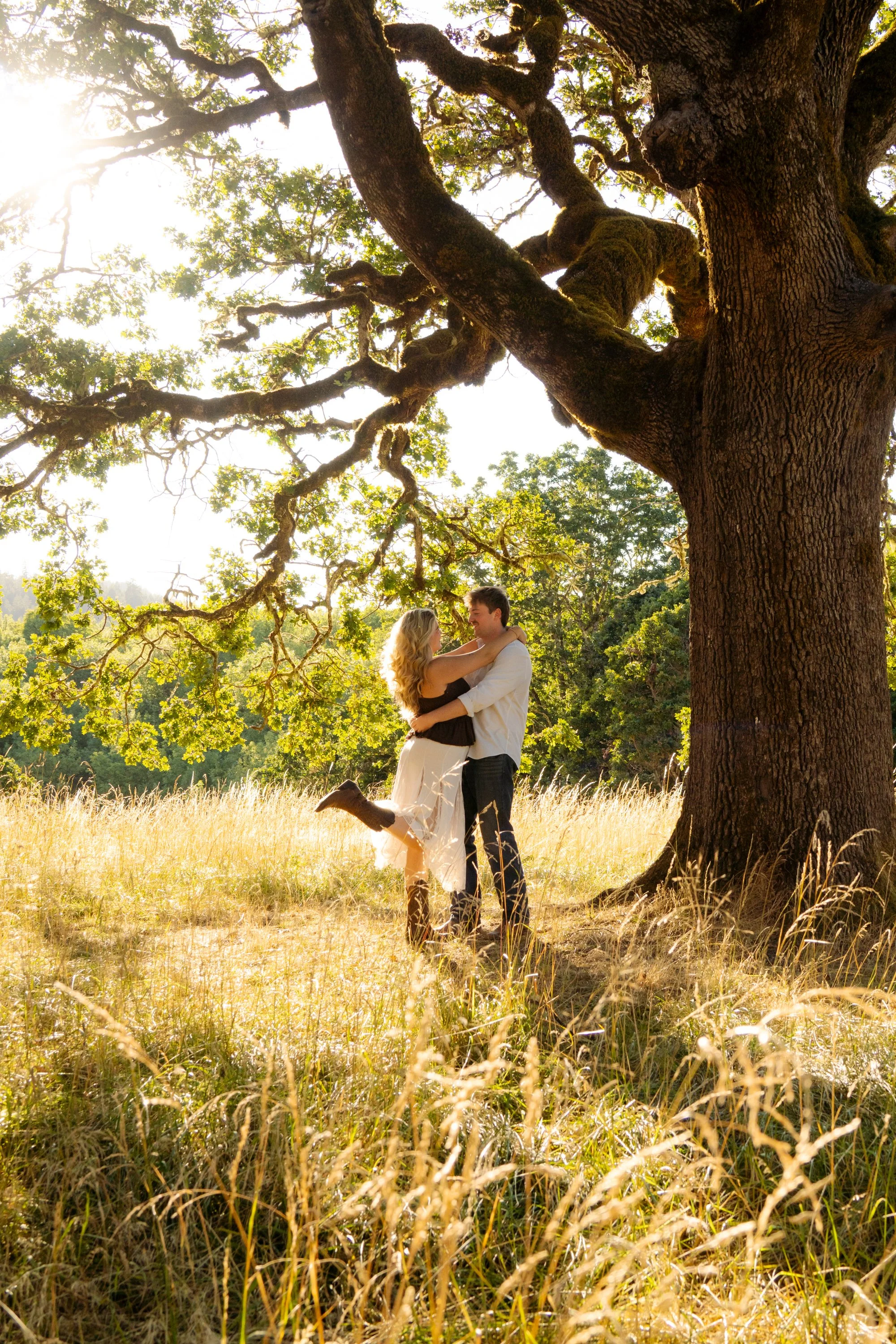 A couple embracing and smiling in a field under a large leafy tree during sunset, with golden light illuminating the scene.