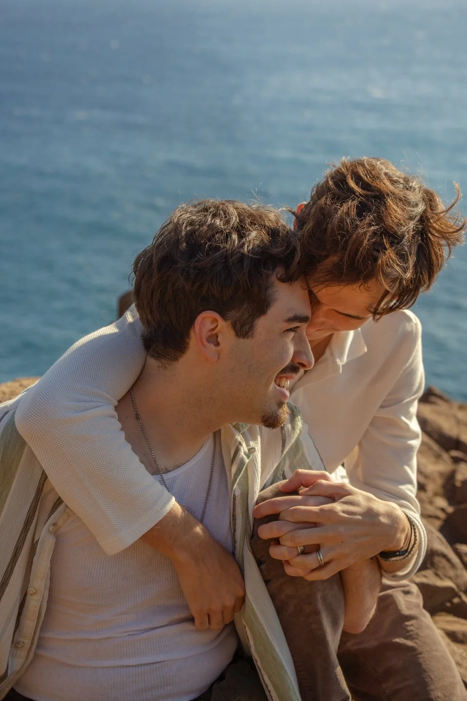 Two people are sitting by the water, smiling and sharing an intimate moment with one person hugging the other from behind, both enjoying a sunny day by the ocean.