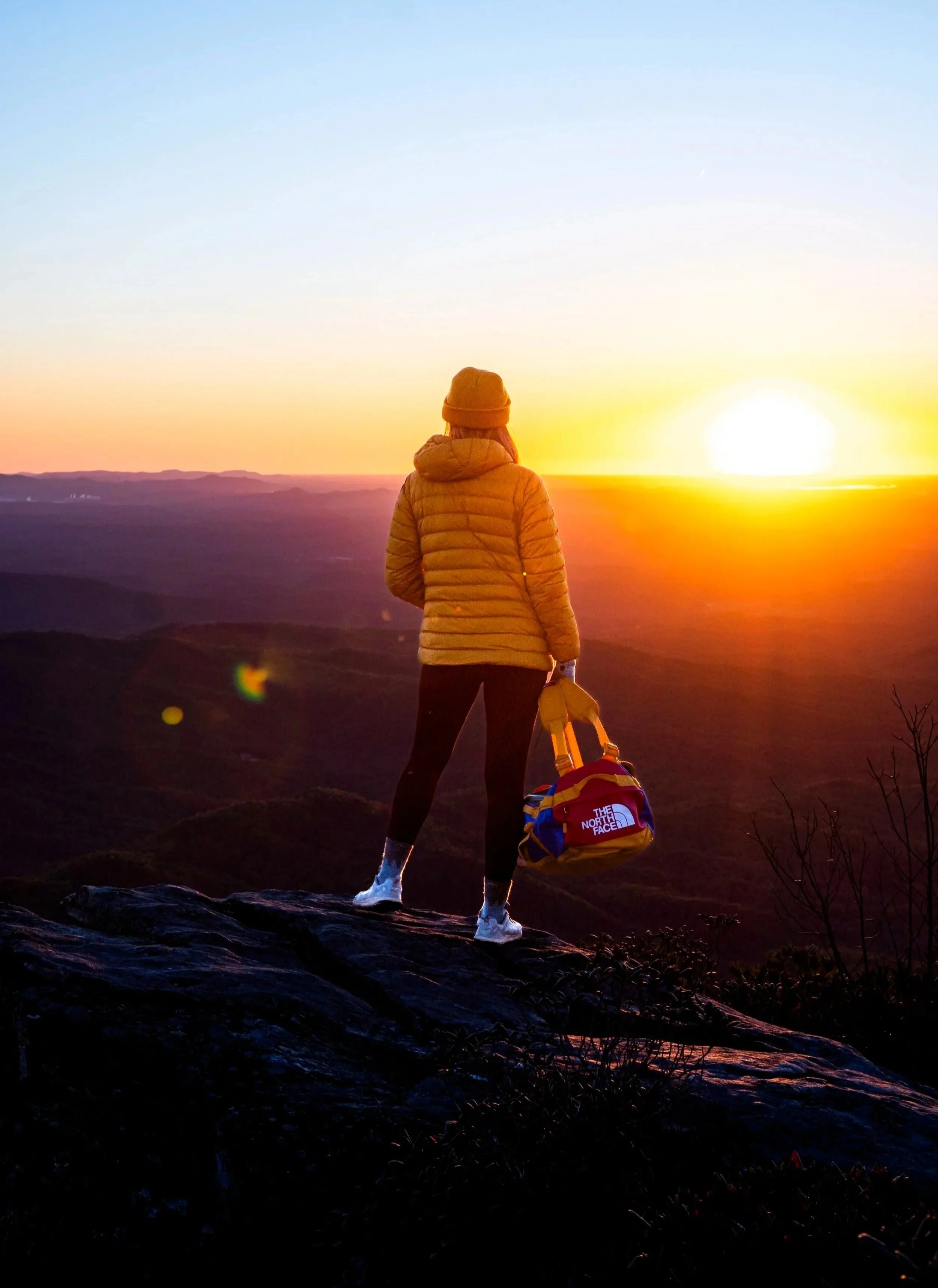 Person wearing a yellow jacket, orange beanie, and black pants standing on a rocky surface, holding a North Face backpack, overlooking a sunset over mountains.