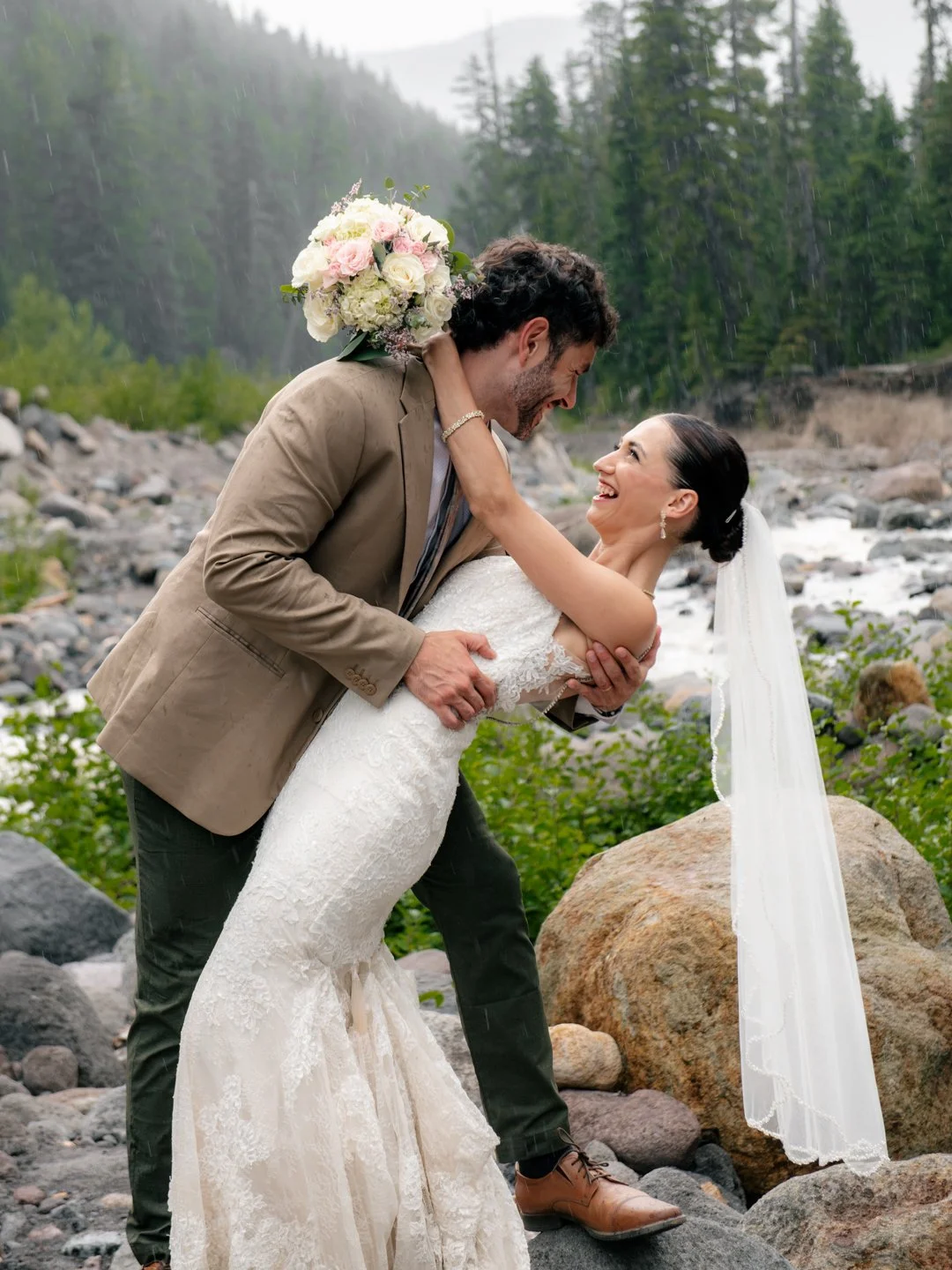 A bride and groom joyful in a forested riverside, with the groom dipping the bride and holding a bouquet of flowers.