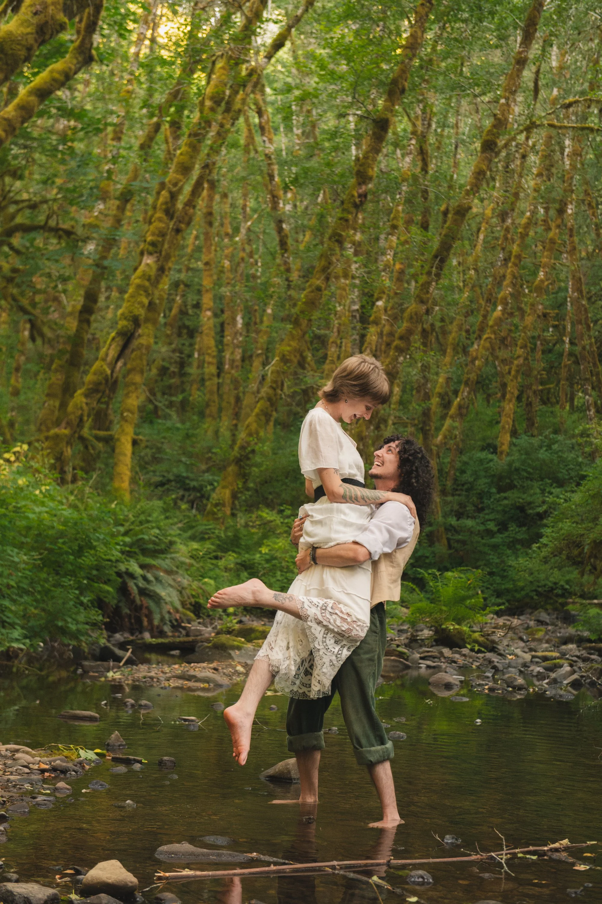 Couple standing in a shallow river surrounded by lush green forest, with the man lifting the woman in his arms. Both are smiling and looking at each other, with the woman barefoot and wearing a white dress with lace details.