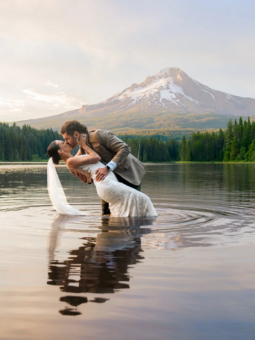 A couple in wedding attire sharing a kiss while standing in a lake with a mountain and forest in the background.