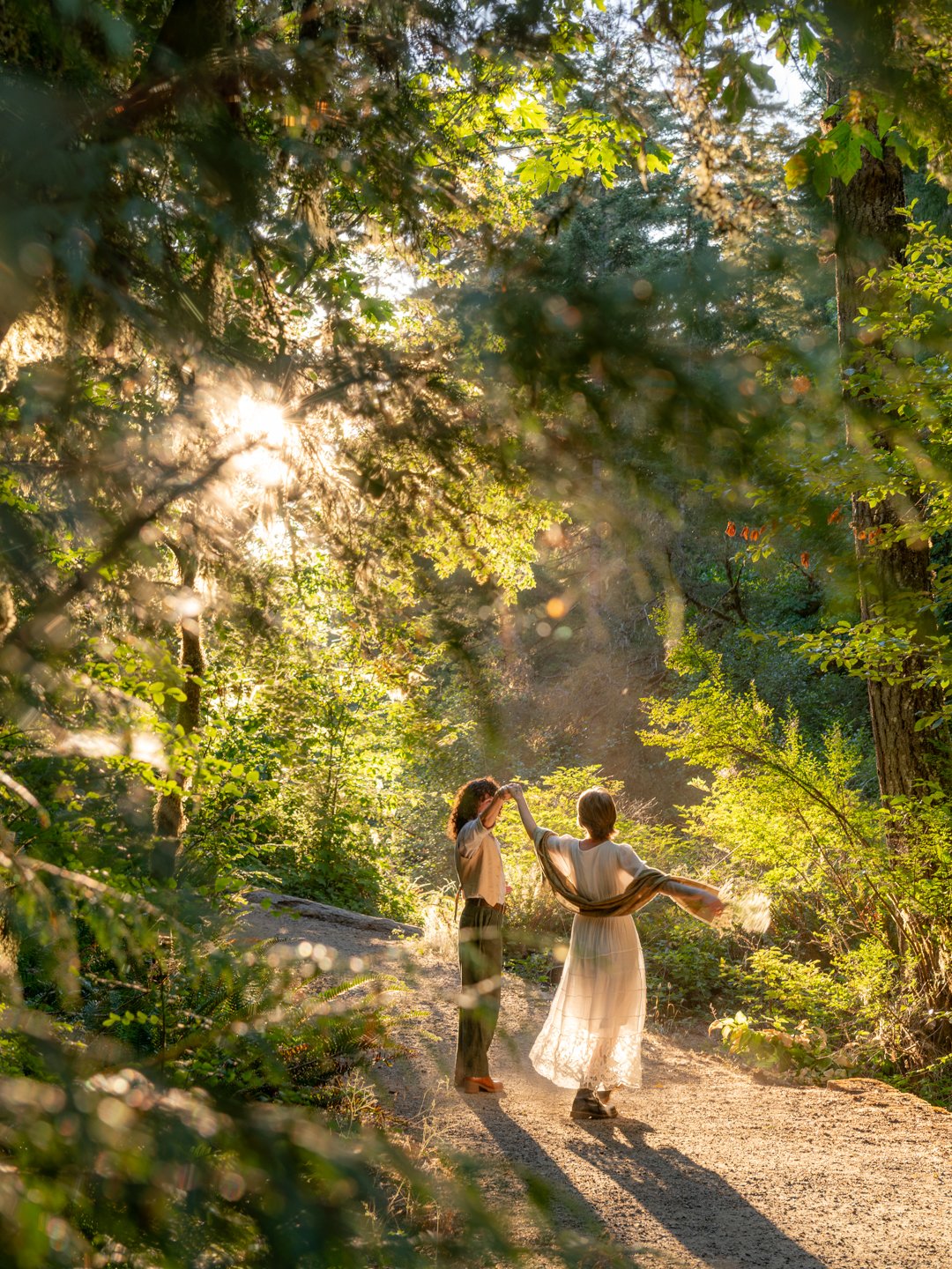 Two people dancing on a forest trail during Golden Hour, surrounded by lush green trees and foliage.